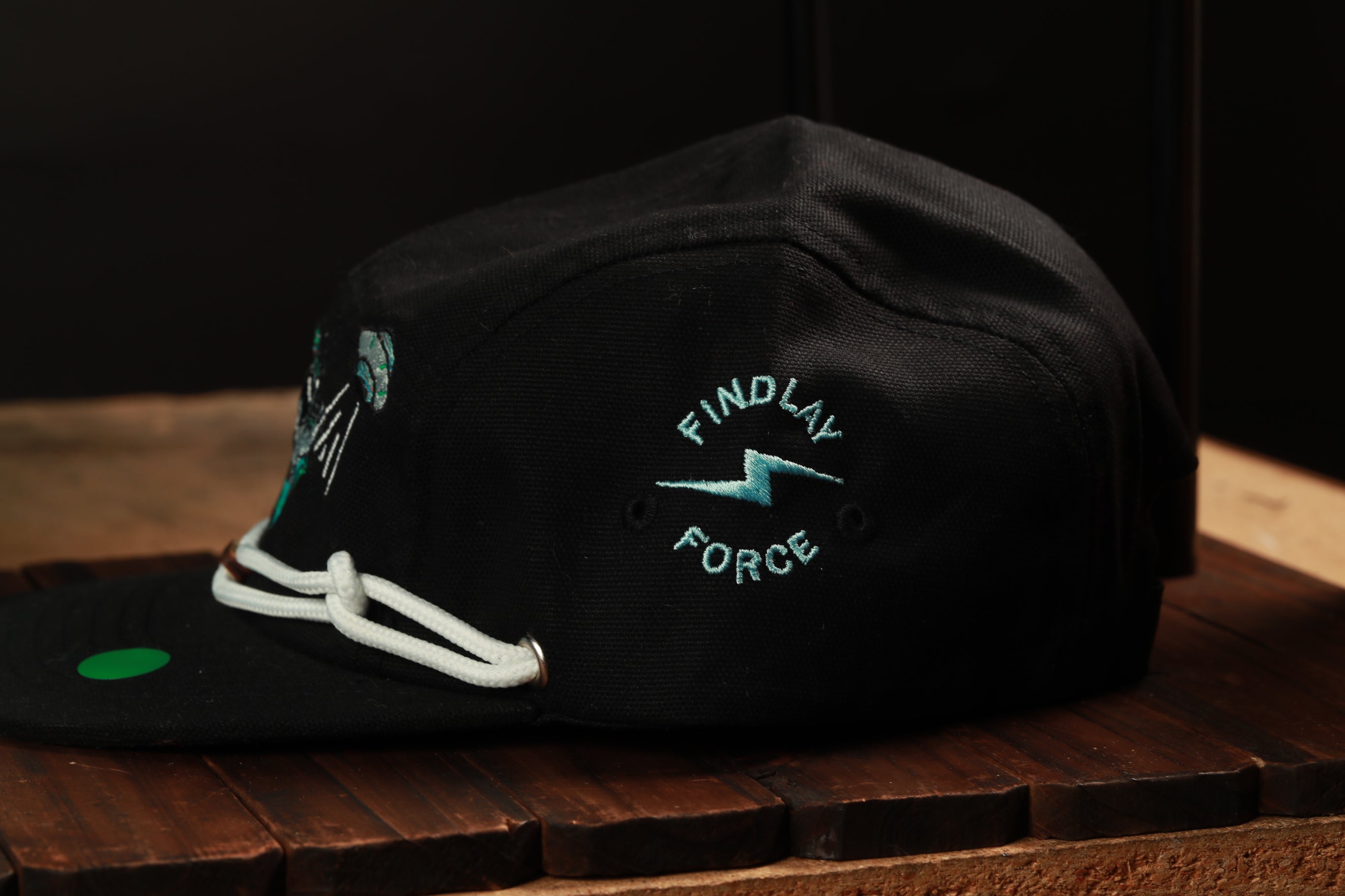 A black baseball cap with a white string and a logo on the front, sitting on a wooden surface.