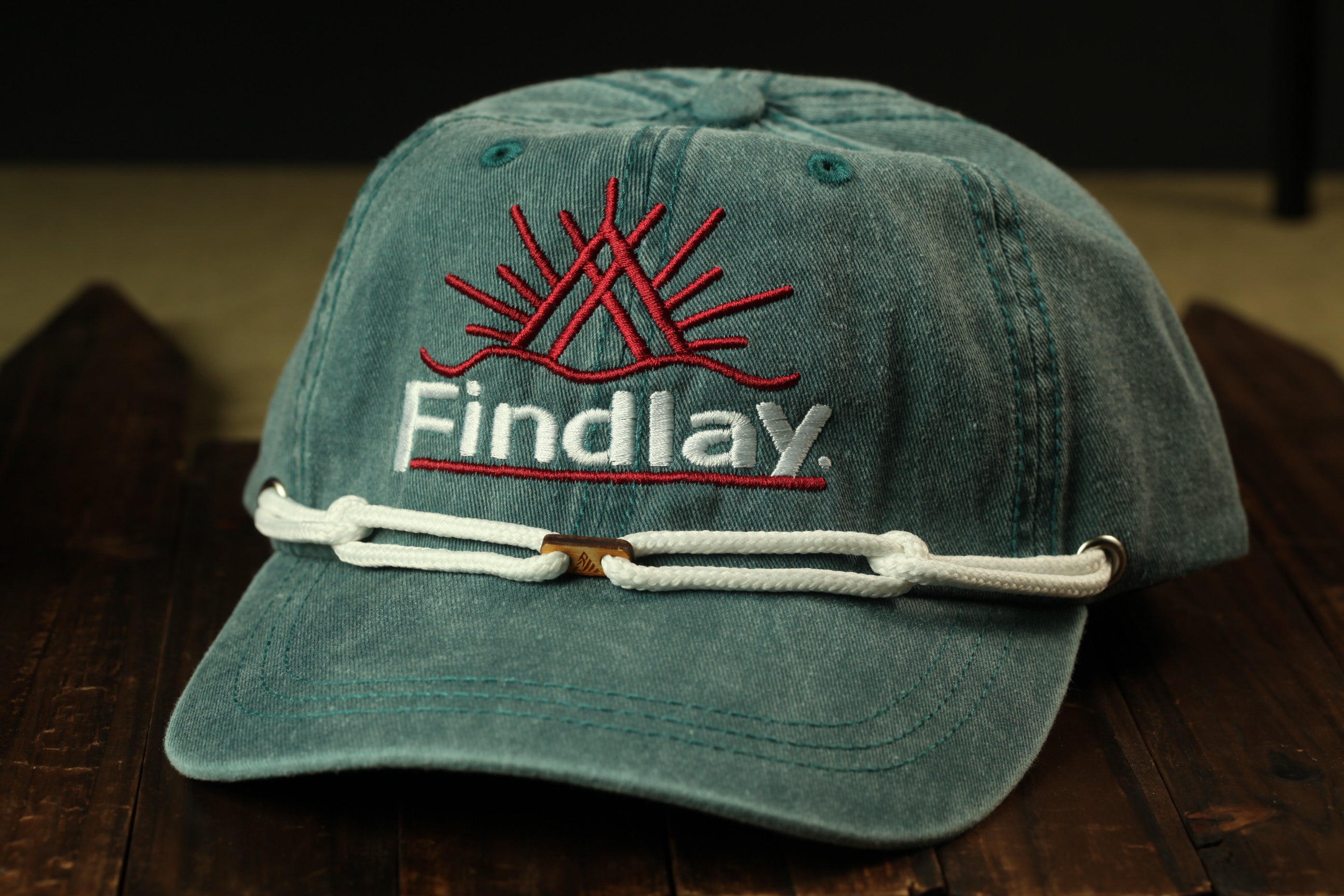 A green baseball cap with a red sun design and the word "Findlay" printed on it, resting on a wooden surface.
