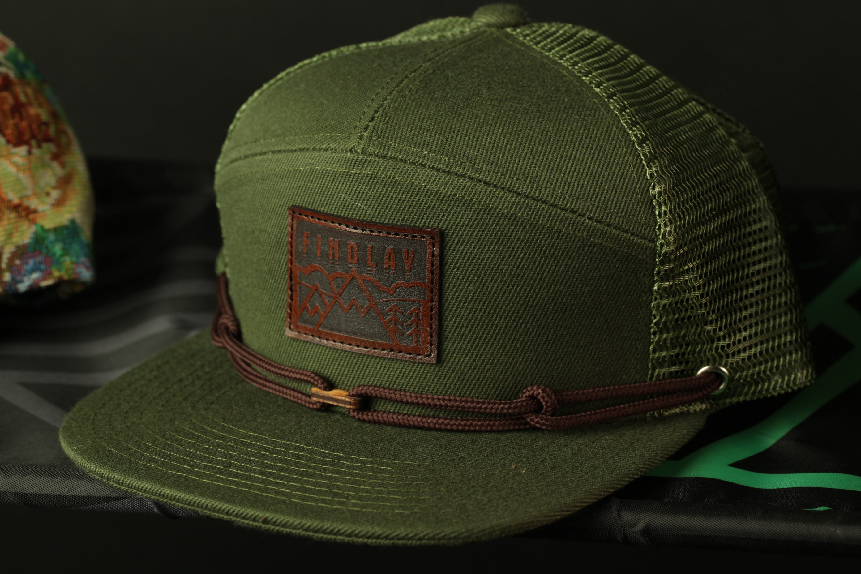 A green baseball cap with a brown leather patch on the front, featuring a mountain design.