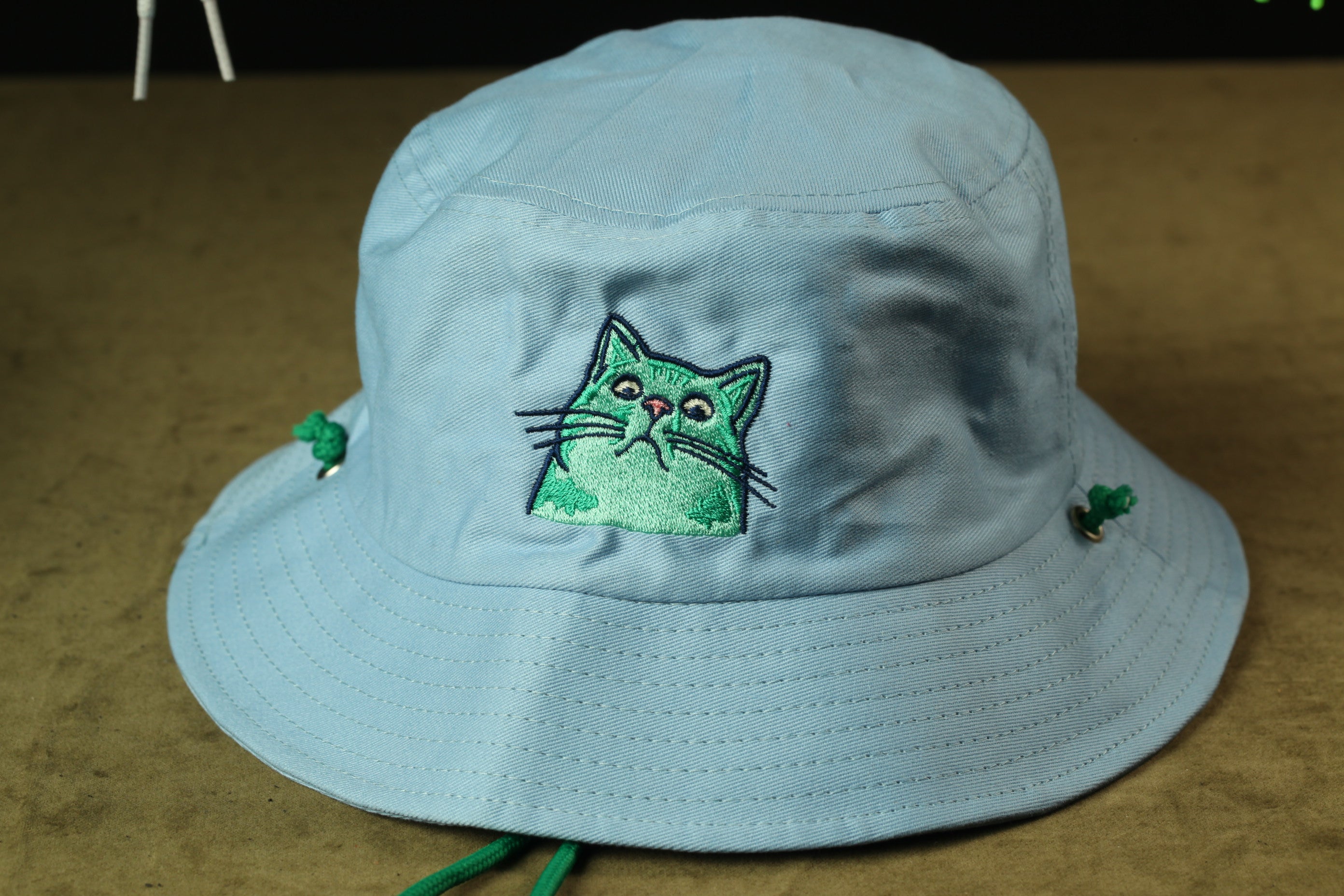 A light blue bucket hat with a green cat embroidered on the front.