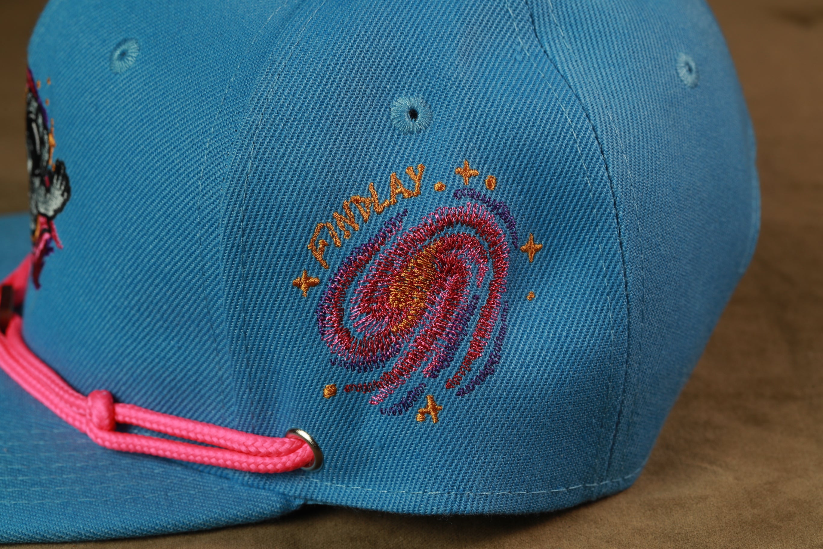 A close-up view of a blue baseball cap with a galaxy embroidered on it, featuring a pink string.