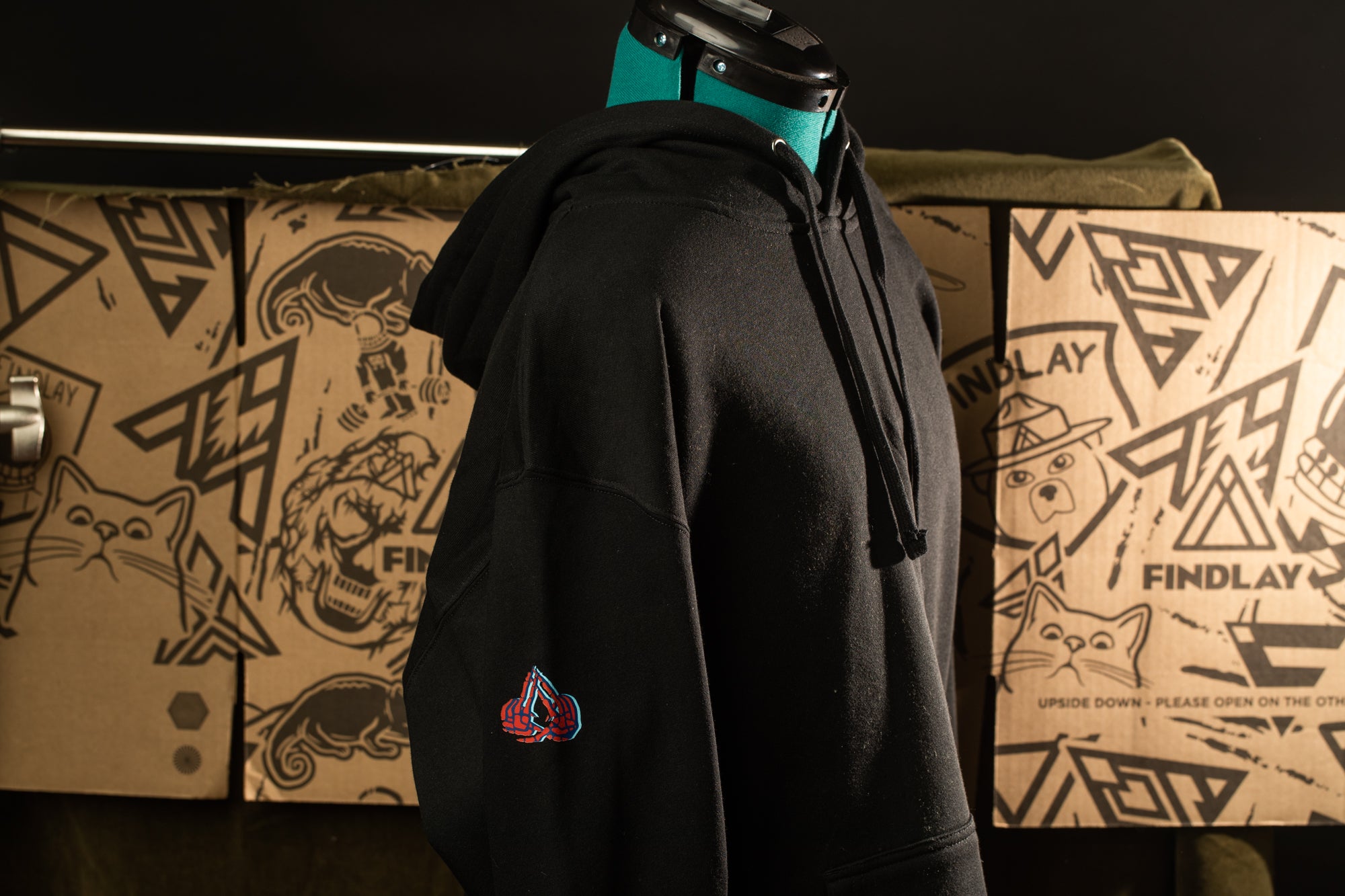 A black hoodie with a teal neckline is displayed on a mannequin, with a cardboard box featuring various graphic designs in the background.