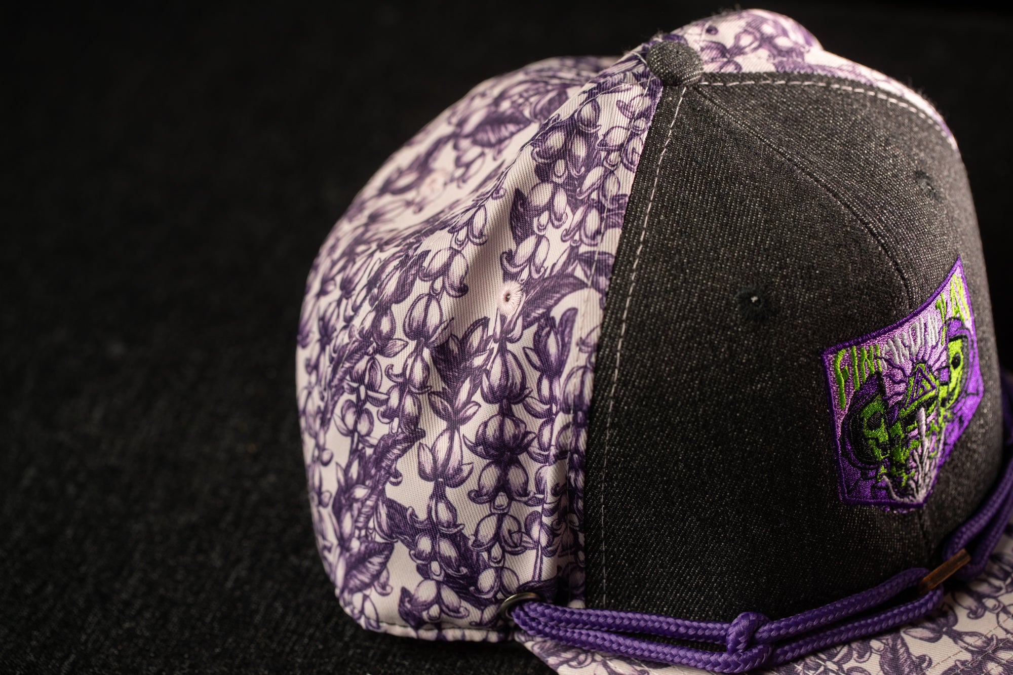 A close-up view of a baseball cap with a floral pattern on the brim and a purple and black color scheme.