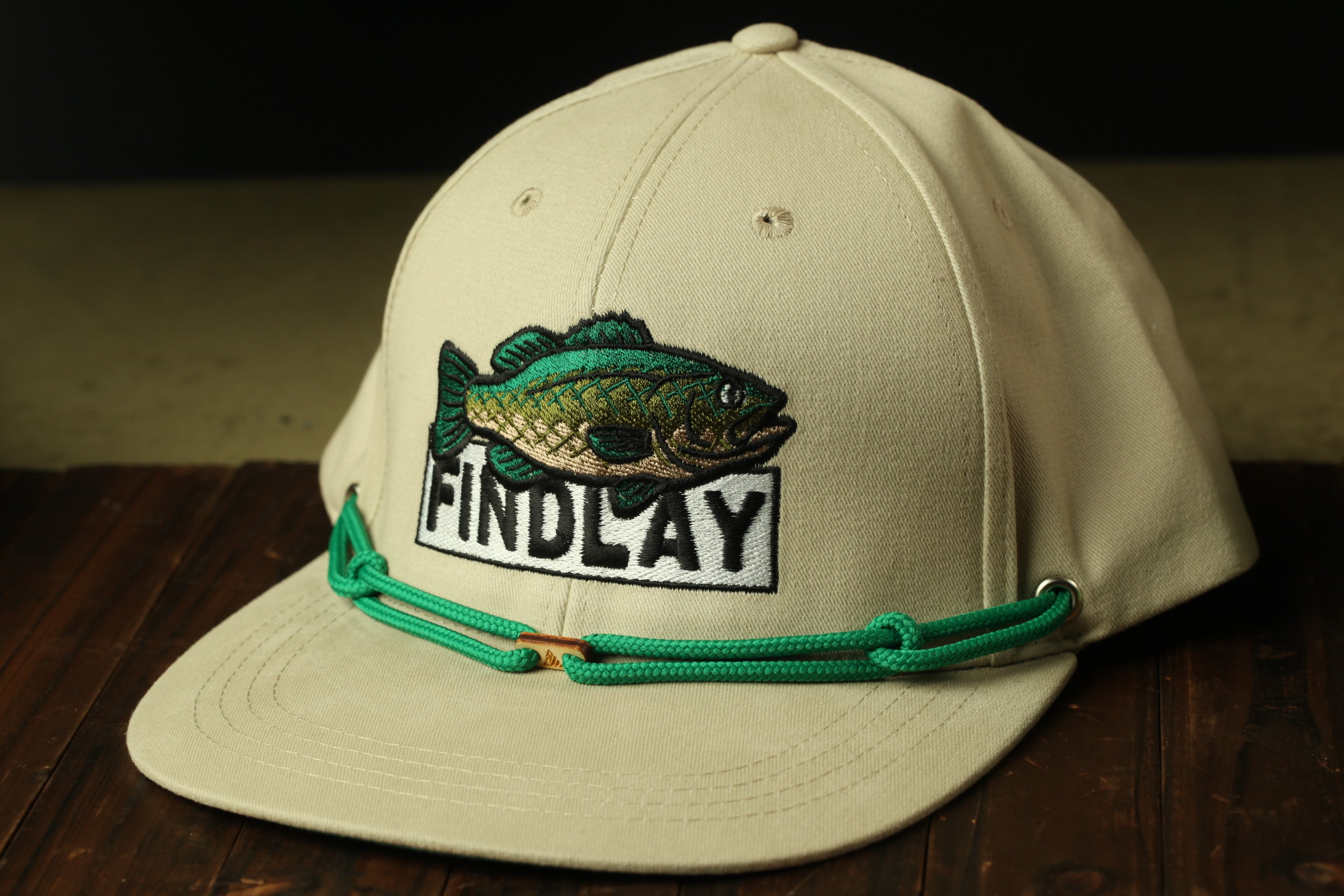 A beige baseball cap with a fish embroidered on it, and the word "FINDLAY" printed on it.