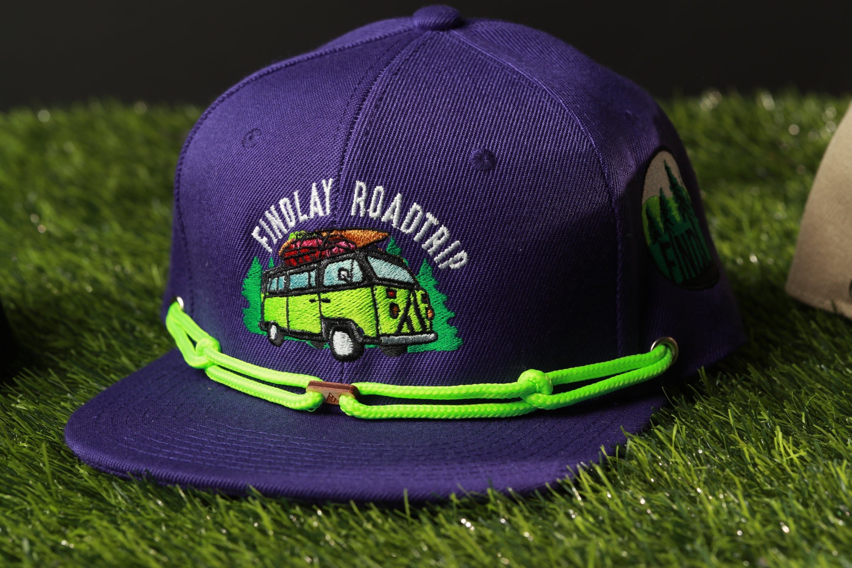 A purple baseball cap with a graphic of a van and the text "Findlay Roadtrip" printed on it, sitting on a grassy surface.
