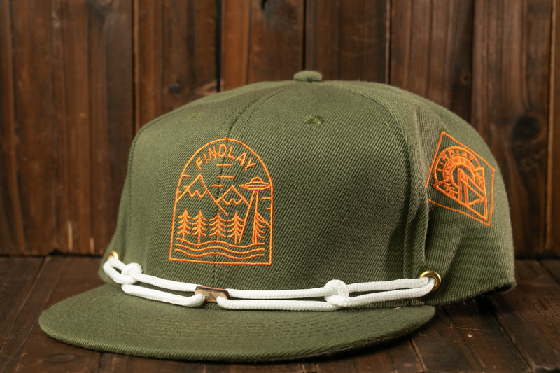 A green baseball cap with a white rope strap and a graphic design on the front, placed on a wooden surface.