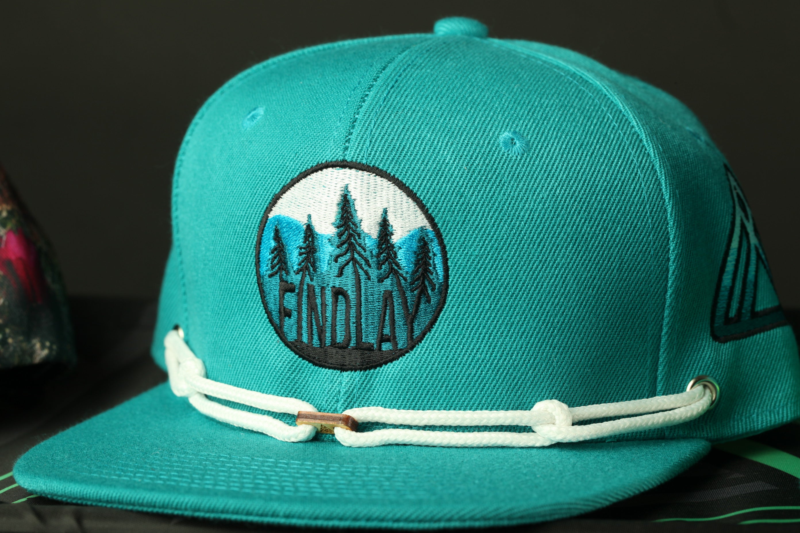 A turquoise baseball cap with a circular embroidered logo on the front.
