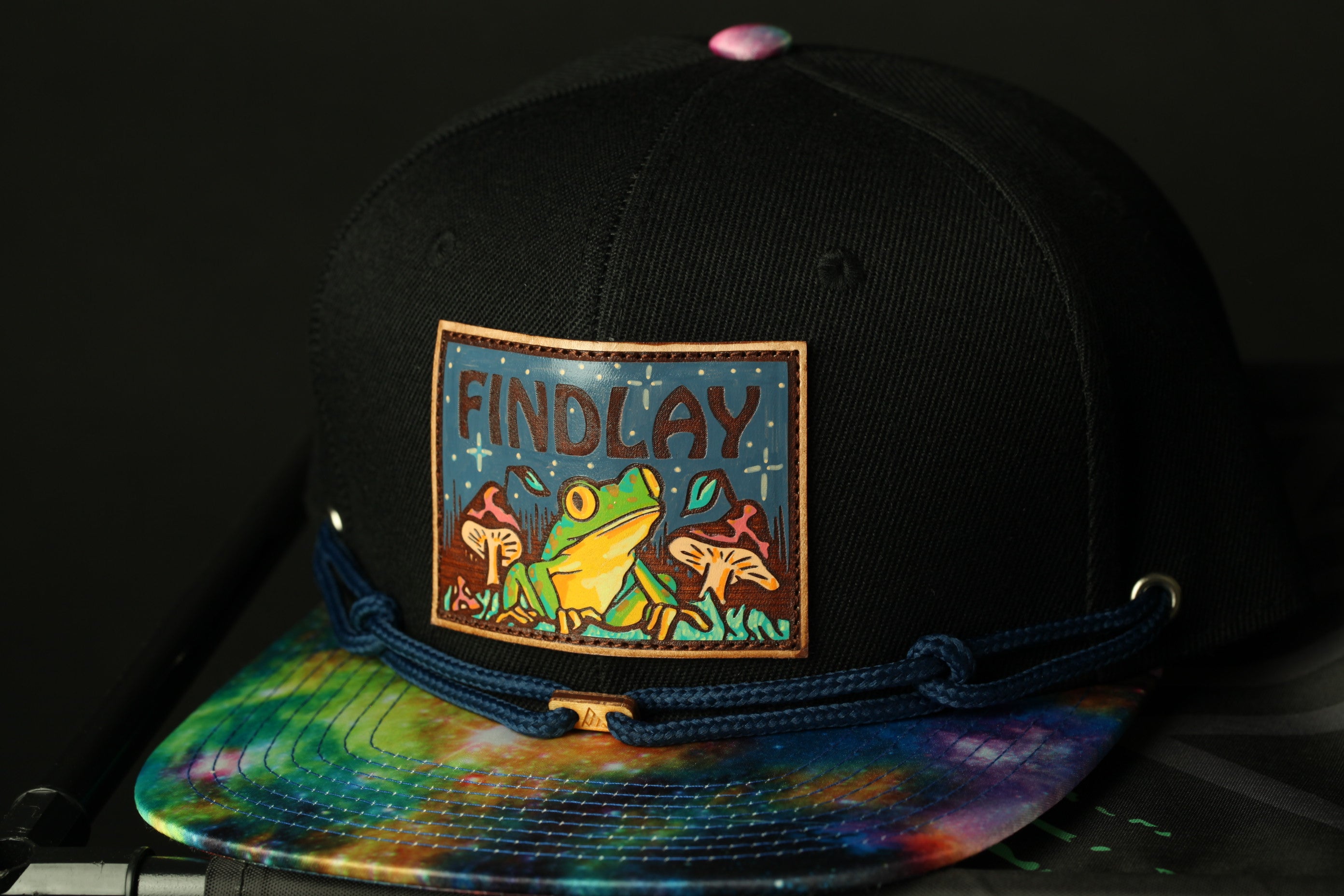 A black baseball cap with a colorful tie-dye pattern and a patch featuring a frog and mushrooms.