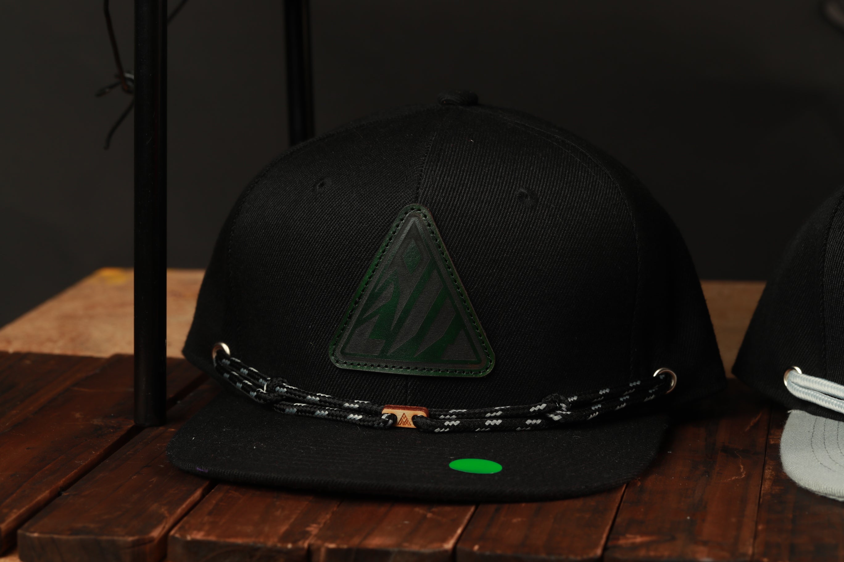 A black baseball cap with a green triangle logo and a black and white braided strap is displayed on a wooden surface.