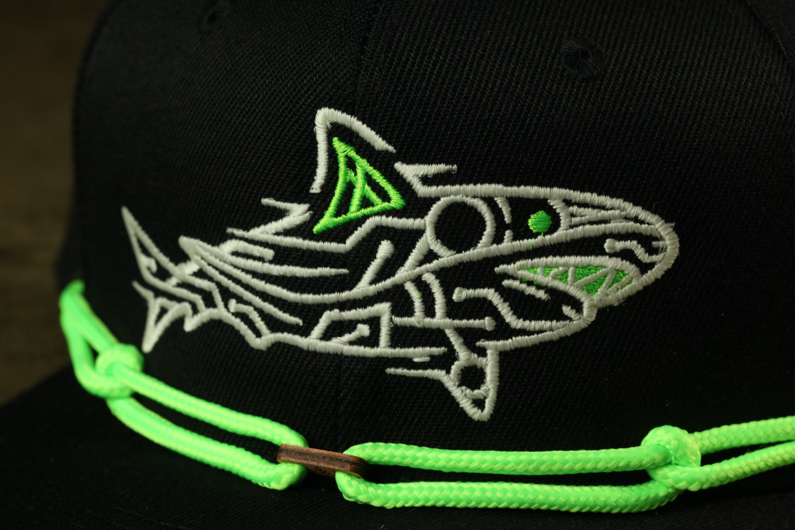 A close-up view of a black baseball cap with a green and white embroidered shark design on the front.