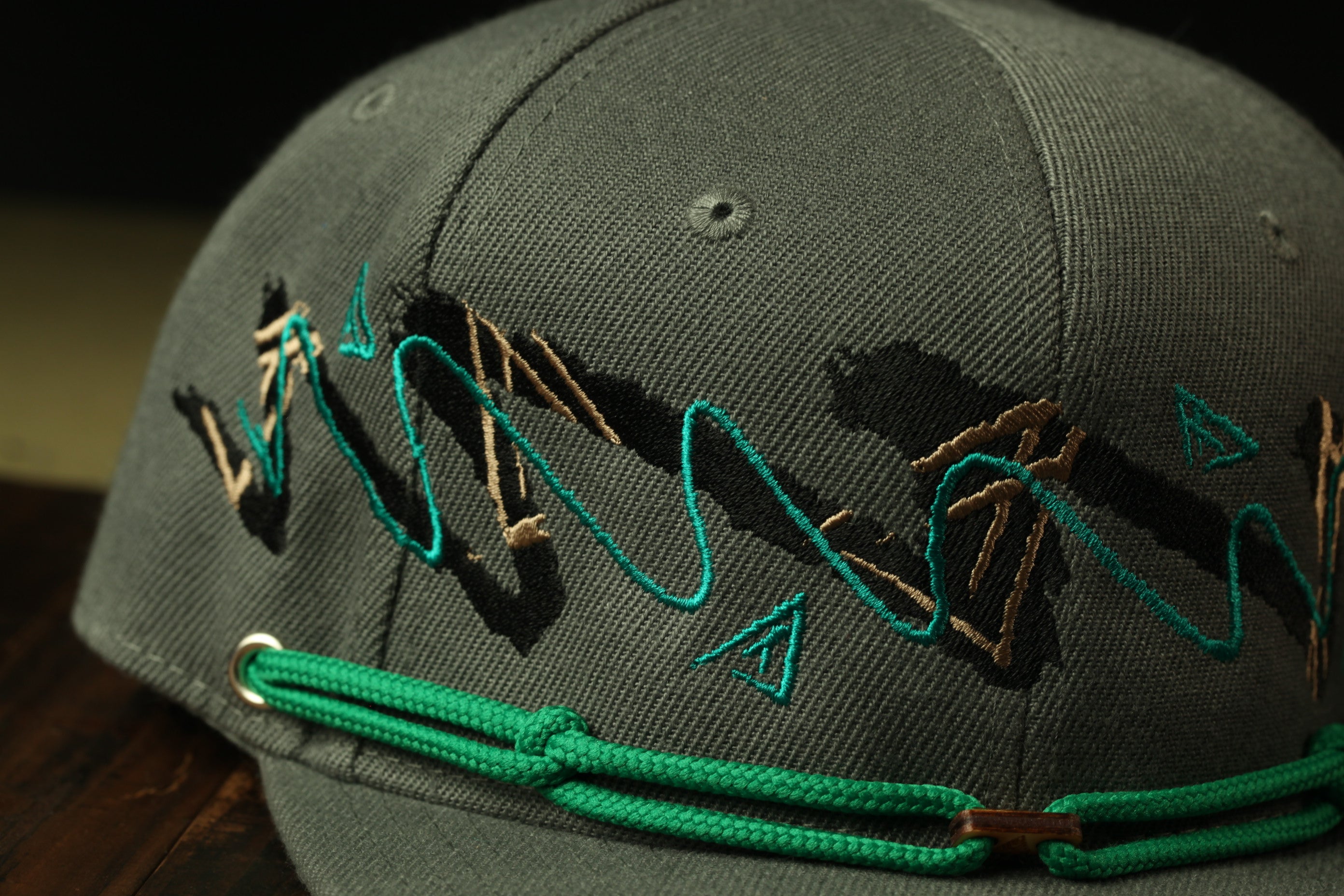 A close-up view of a gray baseball cap with a green string and a pattern of black and gold embroidered designs.