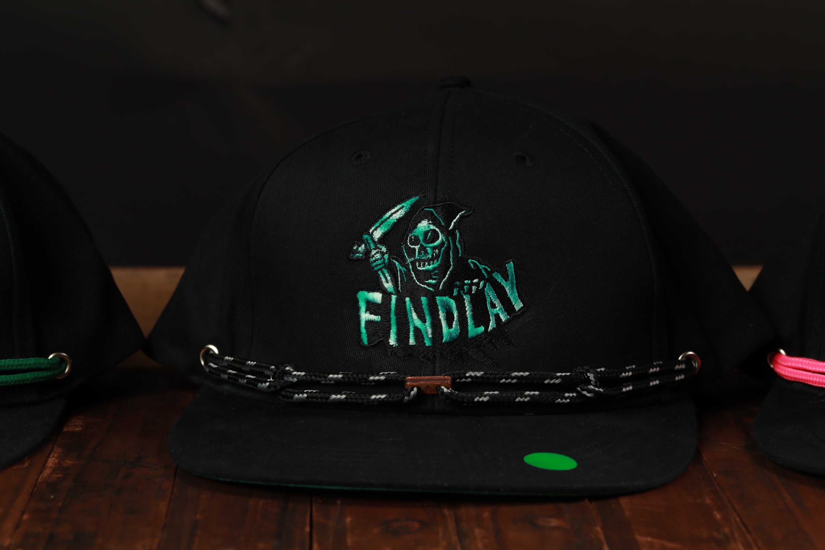 A black baseball cap with a green skull logo and the word "FINDPLAY" printed on it, sitting on a wooden surface.