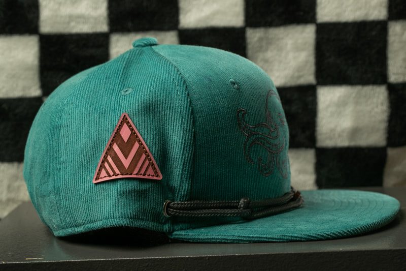A green baseball cap with a triangle patch and an octopus design on the front, placed on a black surface against a checkered background.