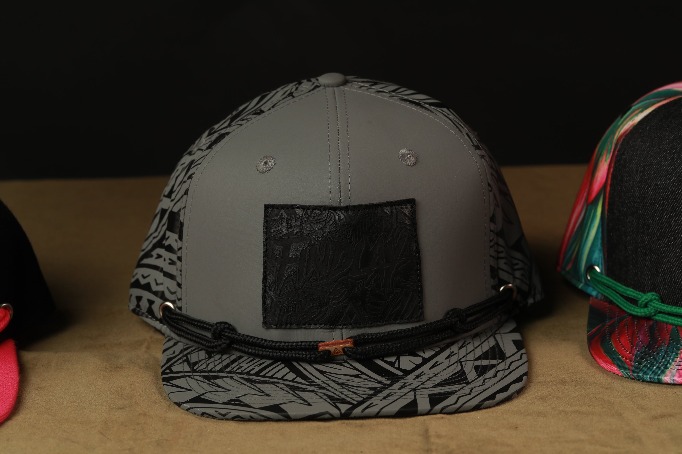 A gray baseball cap with a black patch and a black strap is displayed on a table, along with other colorful baseball caps.