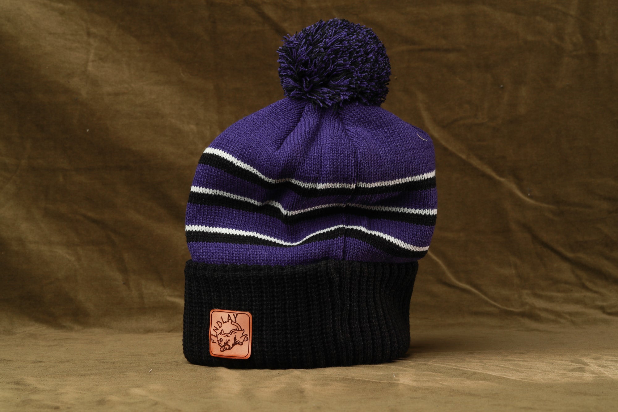 A purple and black striped beanie with a pom pom on top, placed on a brown background.
