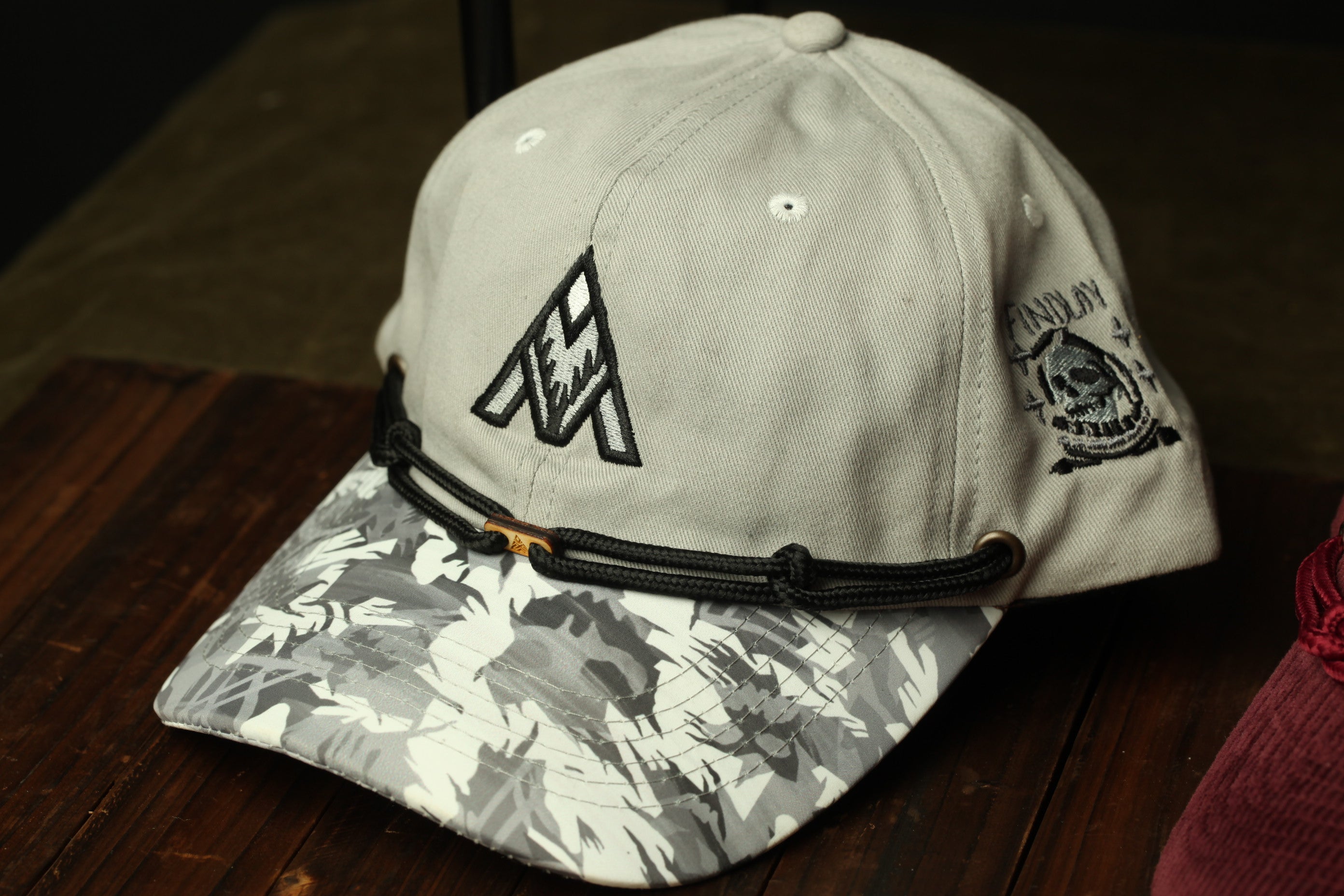 A white baseball cap with a black logo and a camouflage pattern on the brim, placed on a wooden surface.