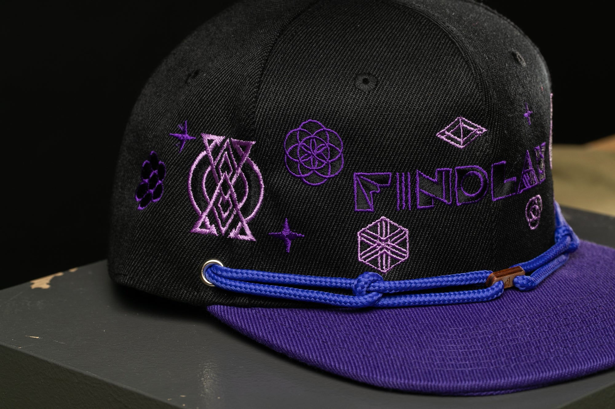A black baseball cap with purple embroidered designs and the word "FINDOLAY" printed on it.