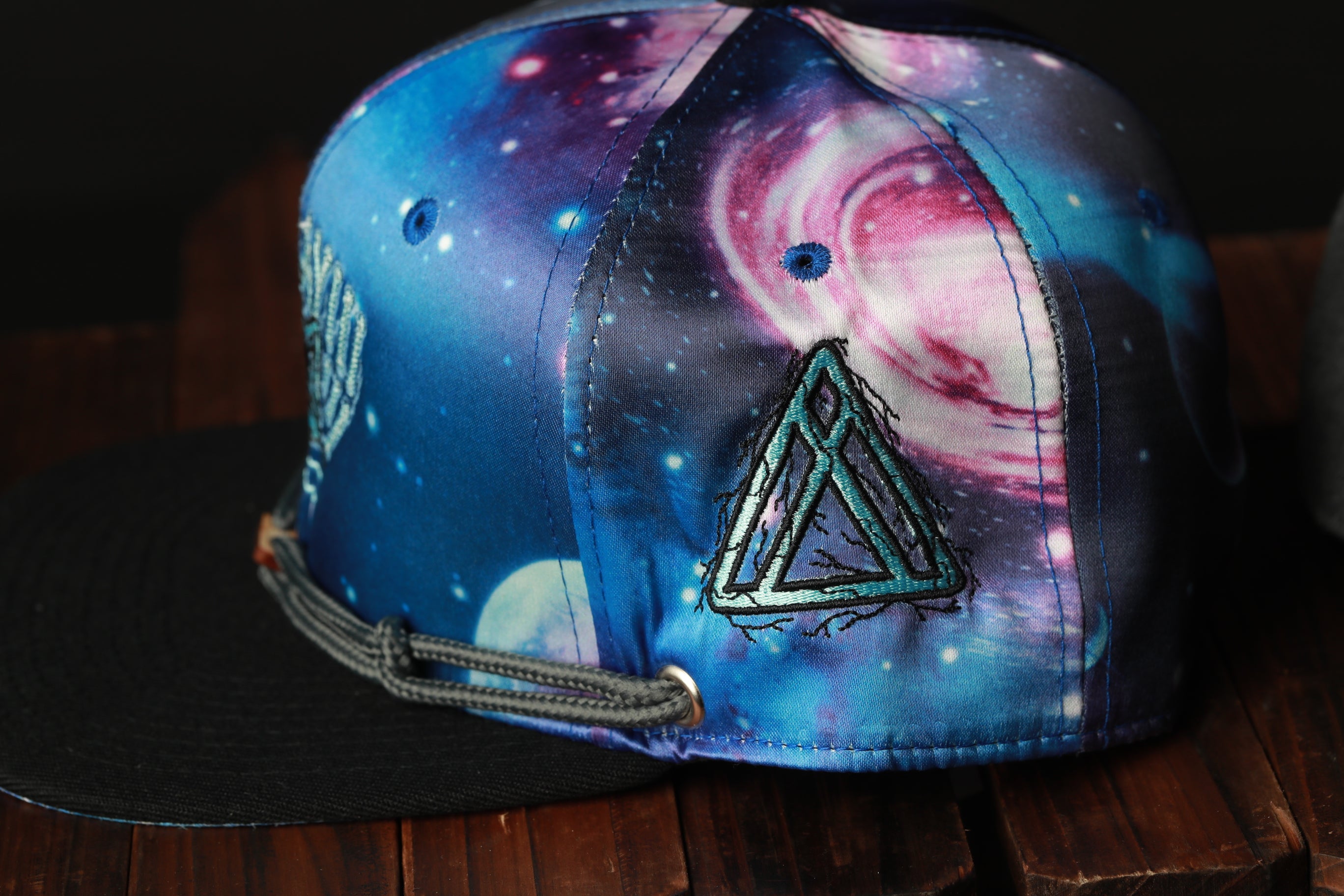 A colorful, patterned baseball cap with a galaxy design and a triangular logo on the front, resting on a wooden surface.
