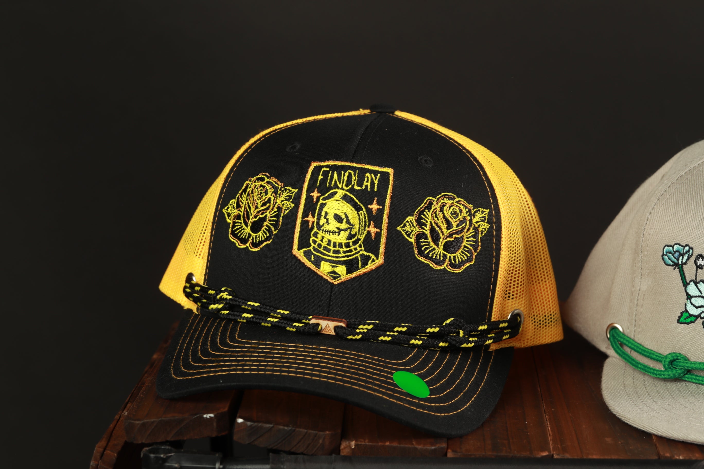 A black and yellow baseball cap with a yellow and black logo, featuring a skull and rose design, is displayed on a wooden surface.