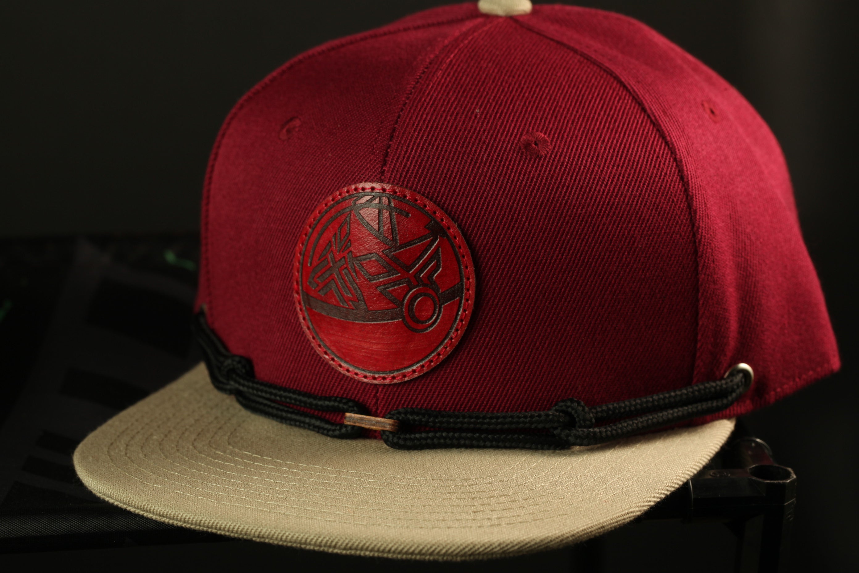 A maroon baseball cap with a red circular logo on the front, featuring a black geometric design.