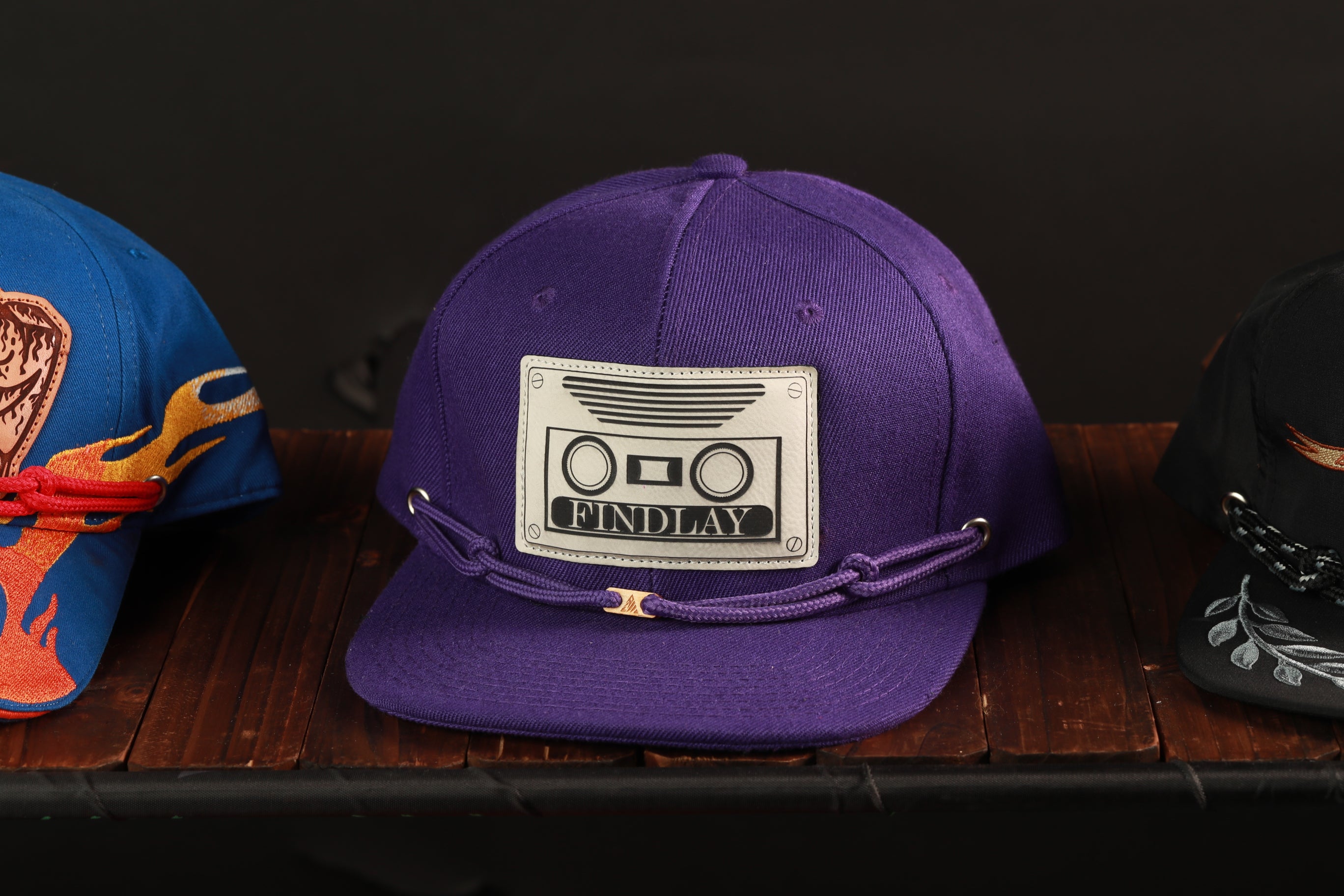 A purple baseball cap with a white label that says "FINDlay" is displayed on a wooden surface, along with other hats.