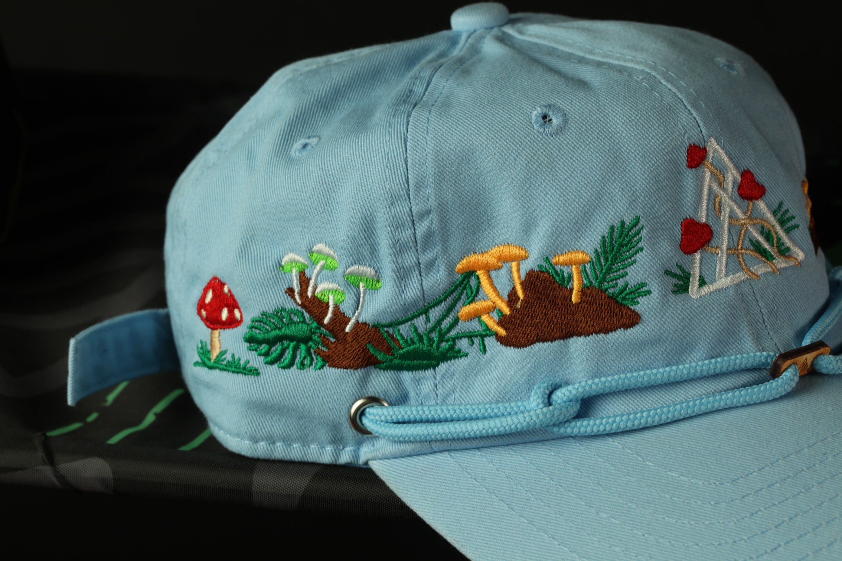 A light blue baseball cap with colorful embroidered designs, including mushrooms, leaves, and flowers, against a dark background.