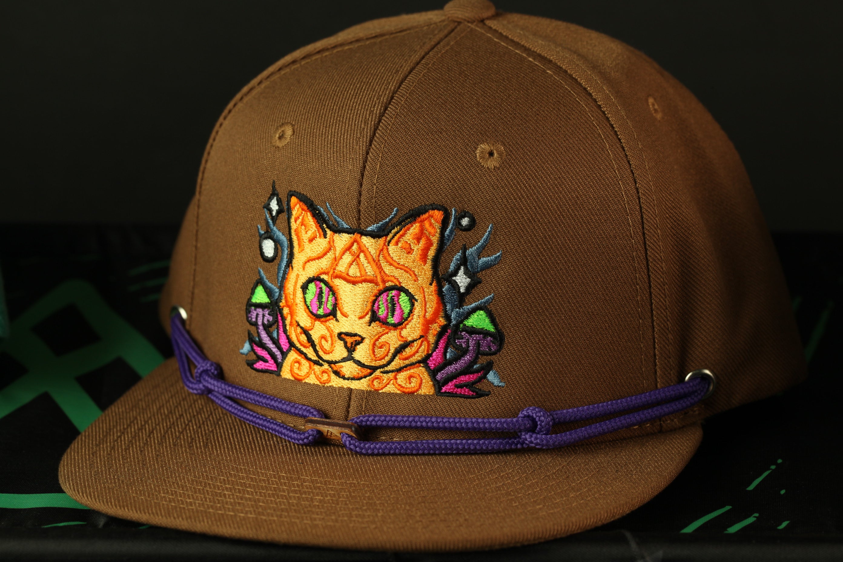 A brown baseball cap with a colorful cat embroidered on it, featuring a purple strap.