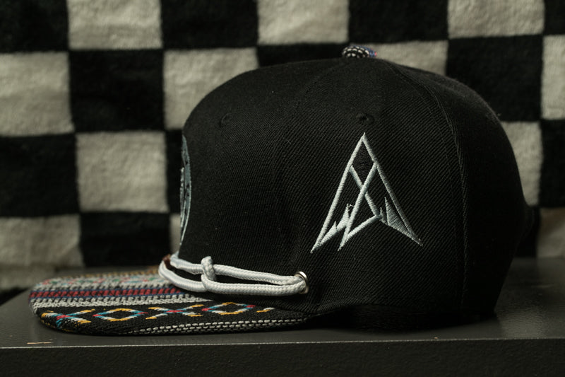 A black baseball cap with a white triangle logo on the front, placed on a patterned fabric surface against a checkered background.