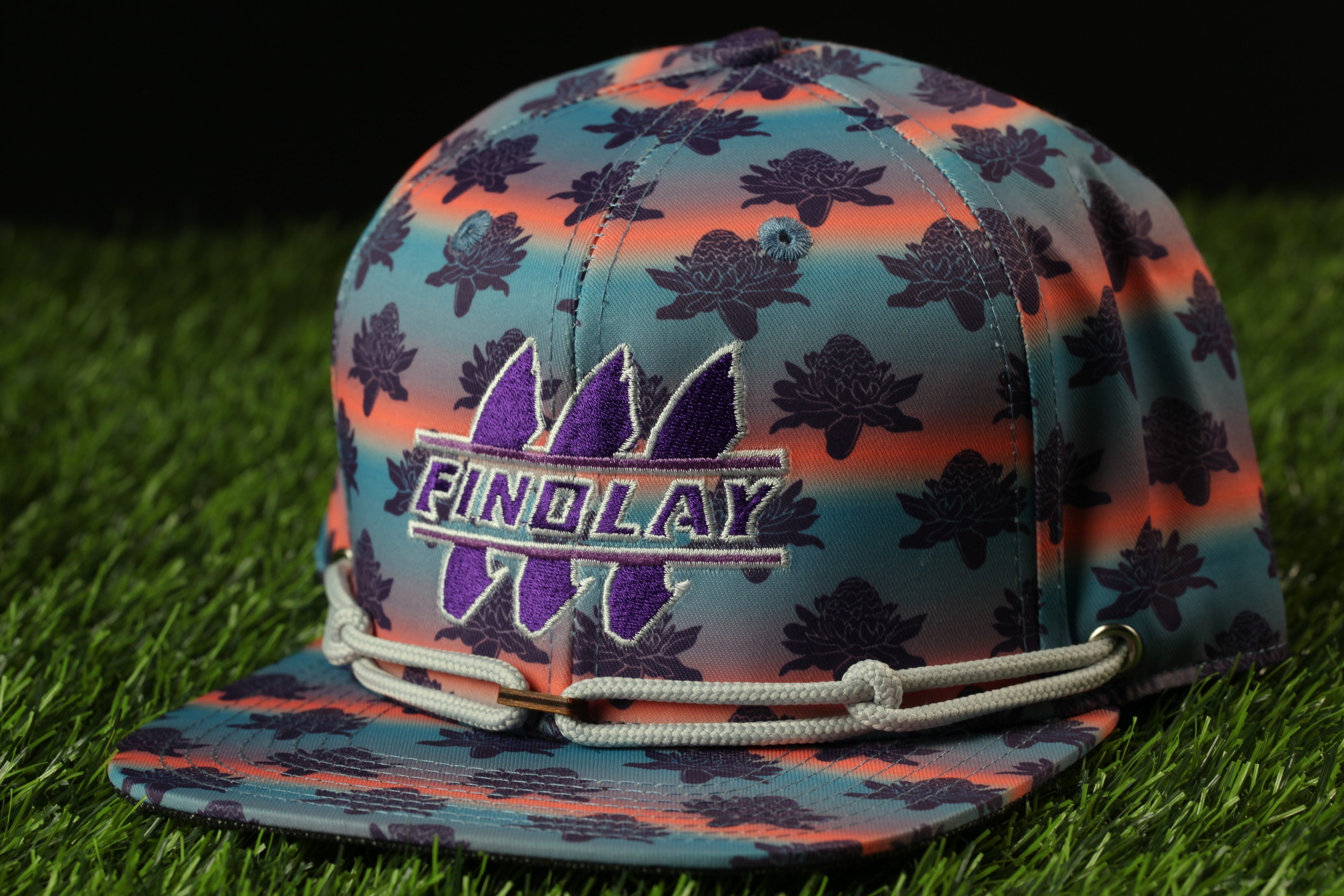 A colorful baseball cap with a floral pattern and the word "FINDOLAY" printed on it, sitting on a grassy surface.