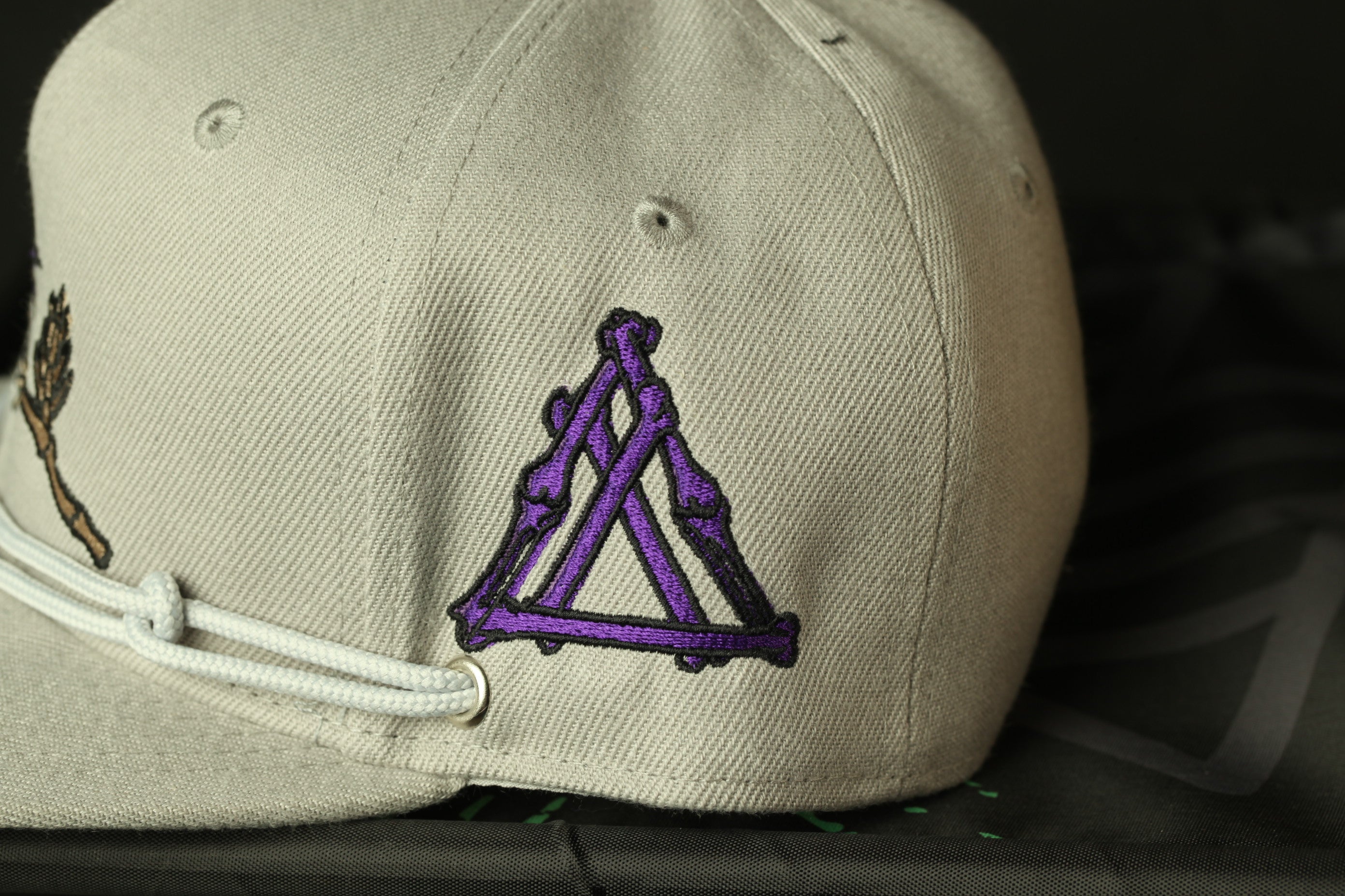 A close-up view of a beige baseball cap with a purple triangle-shaped embroidered patch on the front.