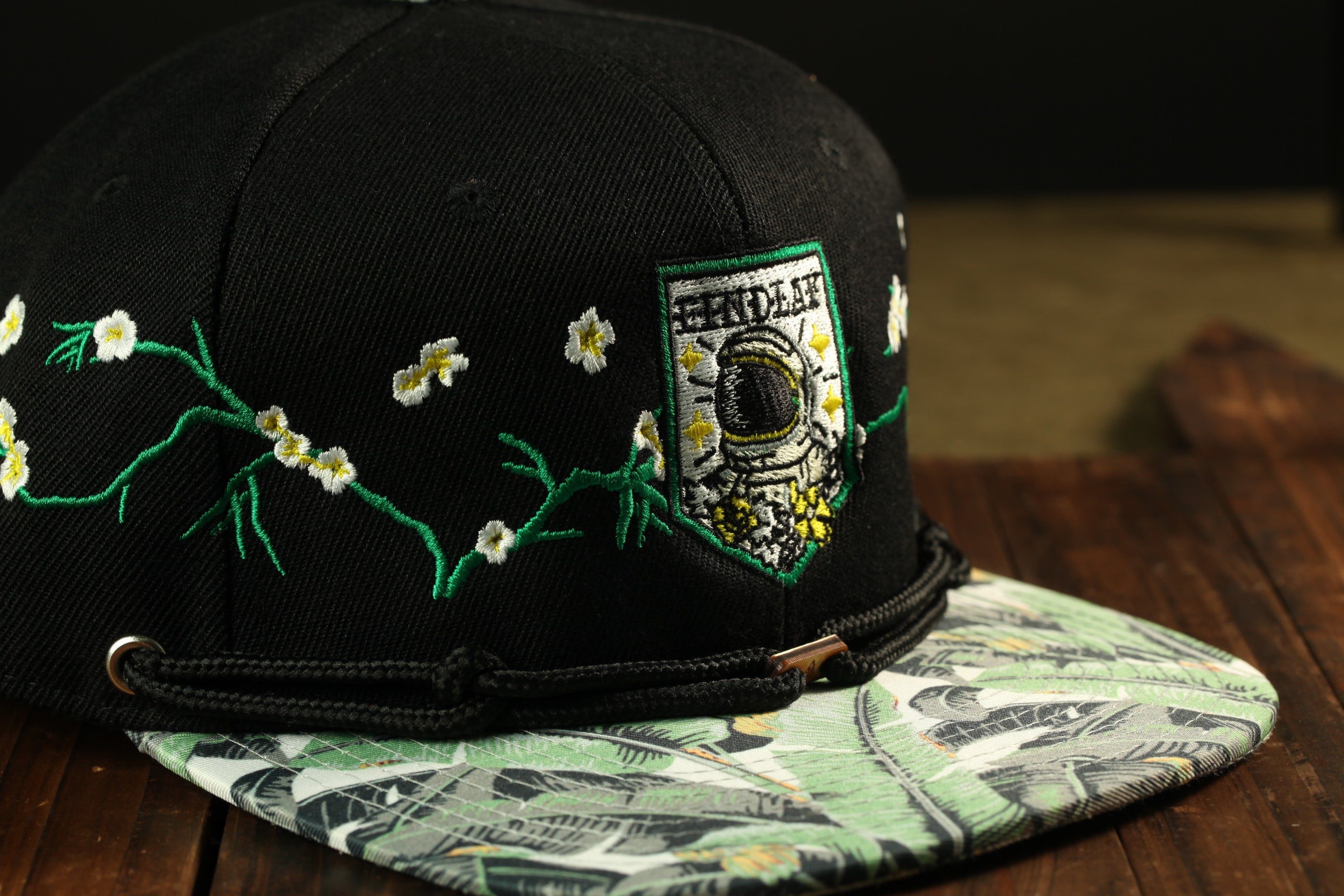 A black baseball cap with a floral pattern and a patch featuring a space suit, placed on a wooden surface.