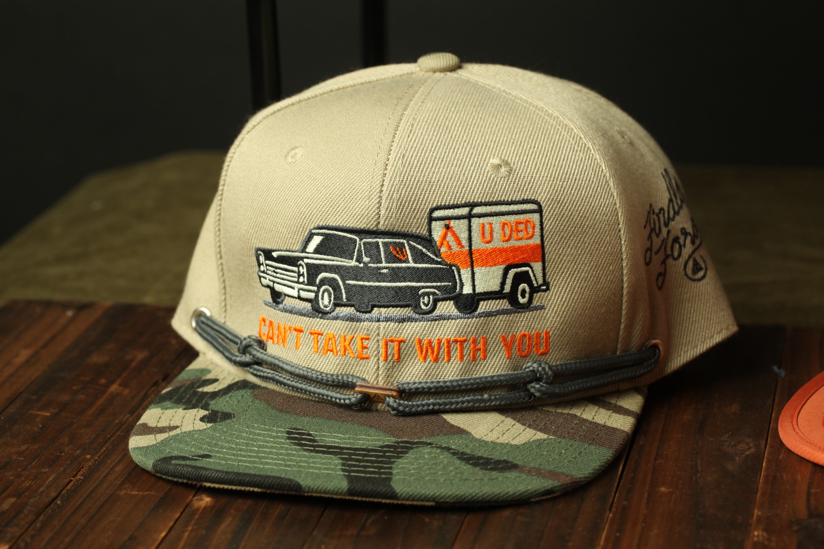 A beige baseball cap with a camouflage pattern and an embroidered image of a car and a trailer.