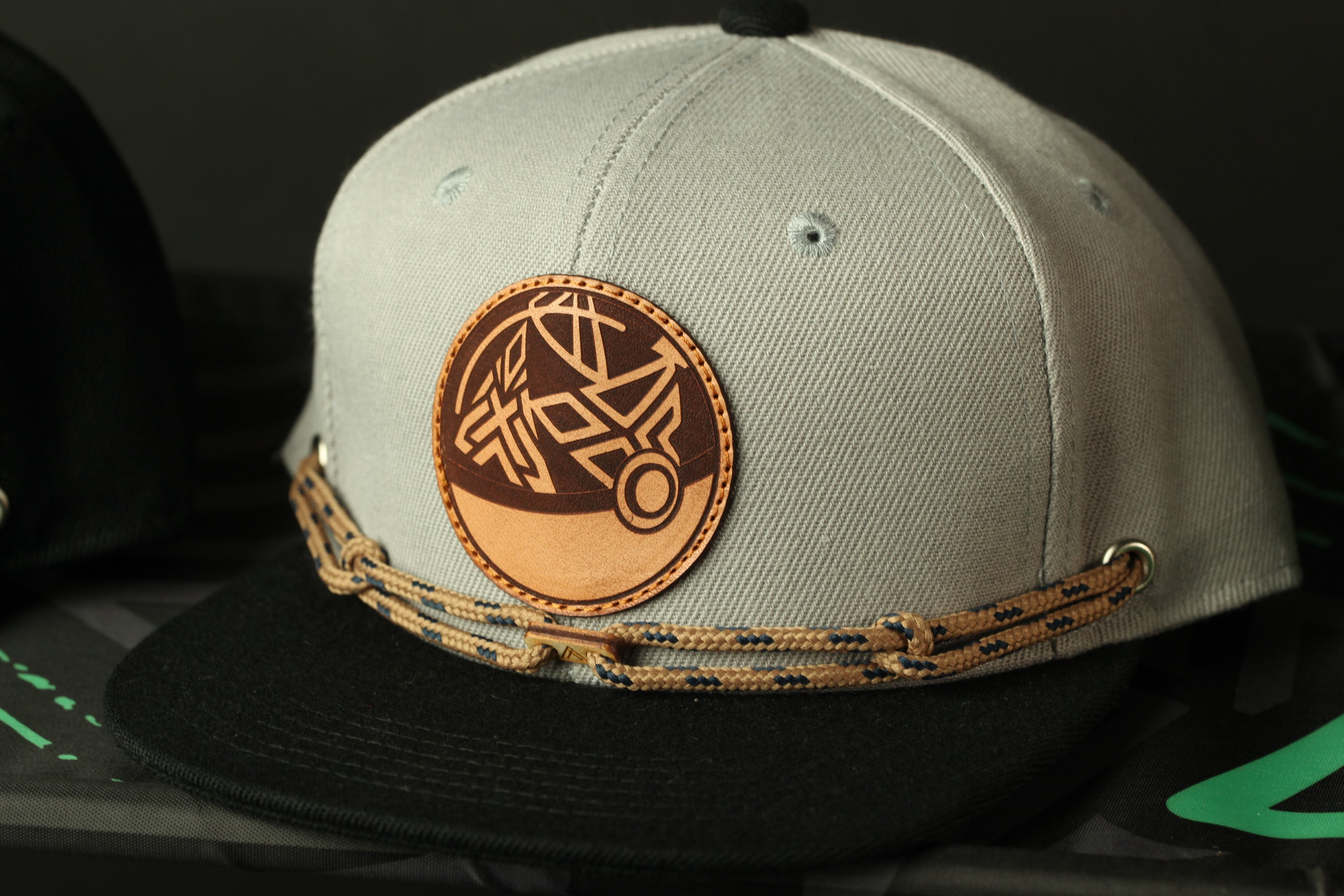 A gray baseball cap with a leather patch and a black brim, featuring a circular logo with a geometric design.