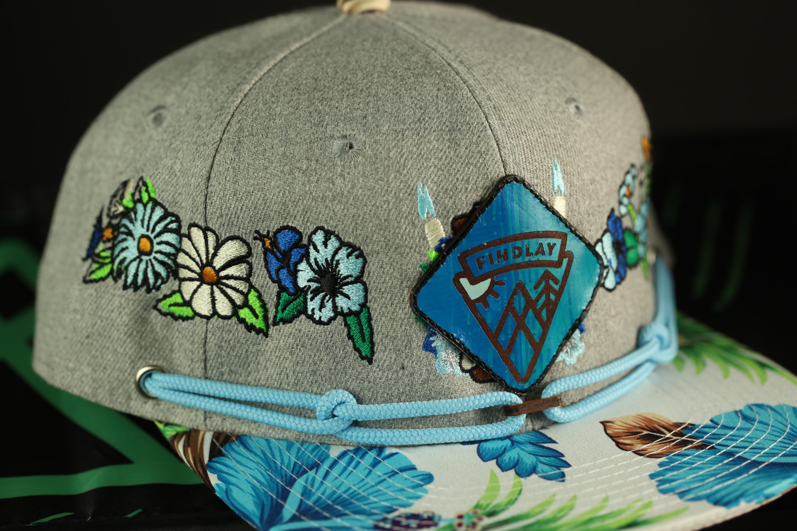 A gray baseball cap with a floral pattern and a patch on the front, featuring a logo.