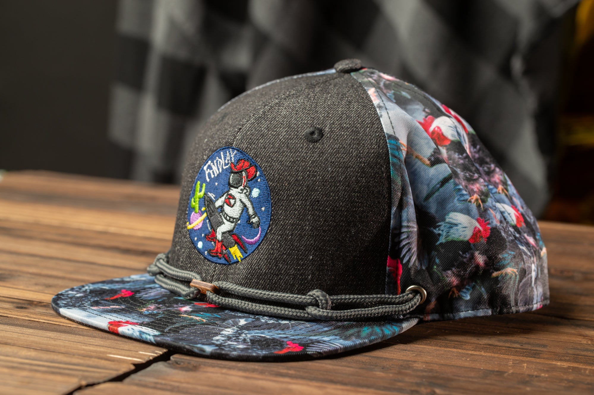 A black baseball cap with a colorful floral pattern and a circular patch featuring a cartoon character is displayed on a wooden surface.