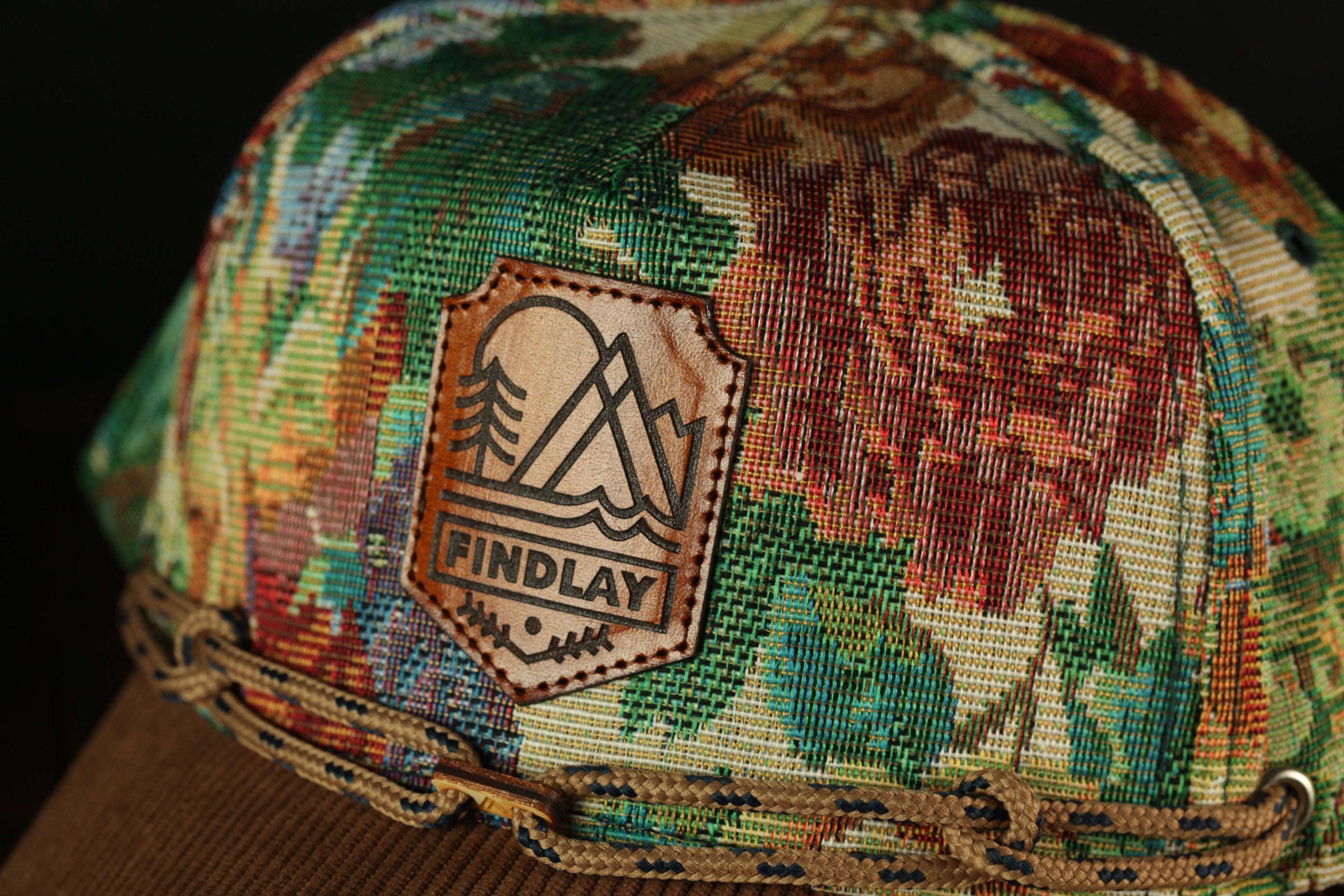 A close-up of a colorful, patterned baseball cap with a leather patch on the front displaying the word "FIND lay".