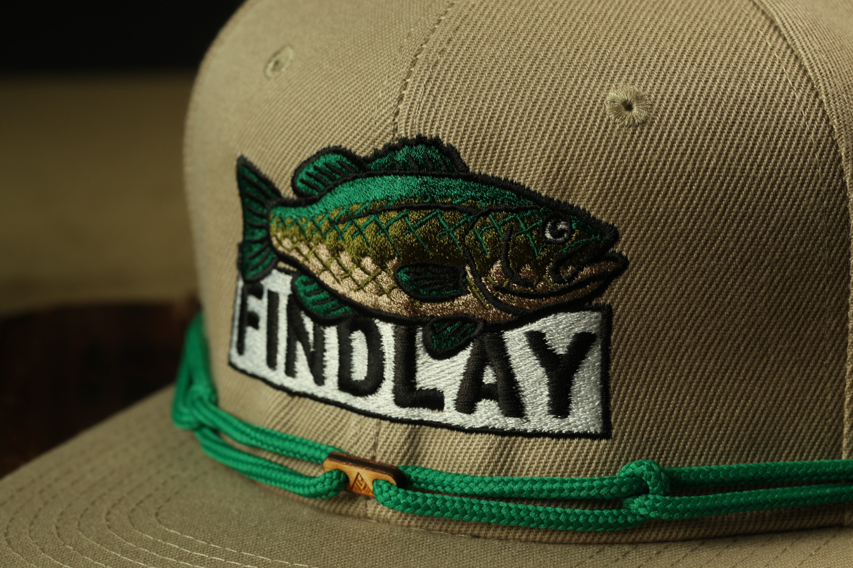 A close-up view of a tan baseball cap with a green string and a fish embroidered on it.