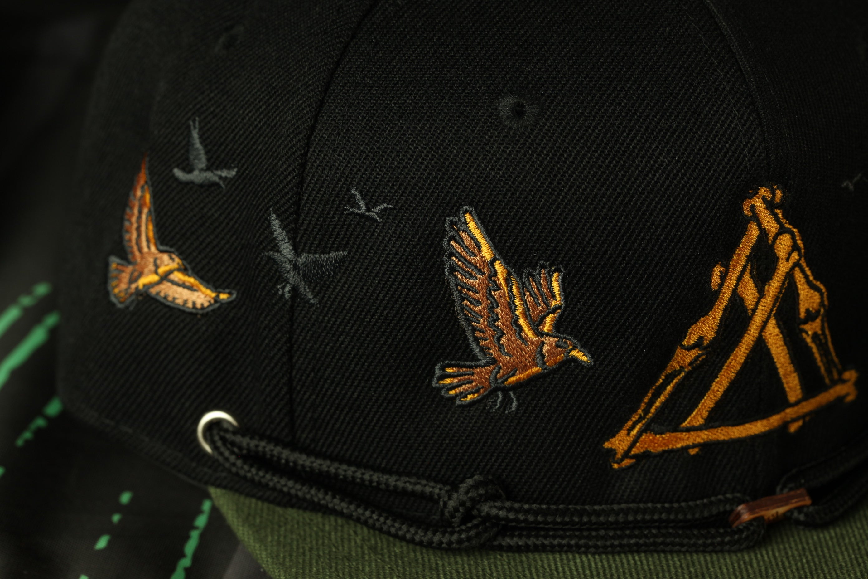 A close-up view of a black baseball cap with gold and orange embroidered designs, including a bird and a triangle.