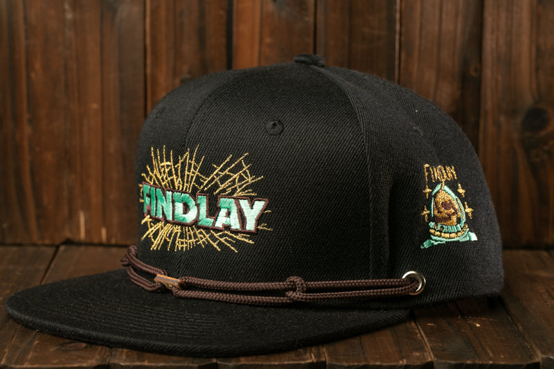 A black baseball cap with a gold embroidered logo and text on it, placed on a wooden surface.