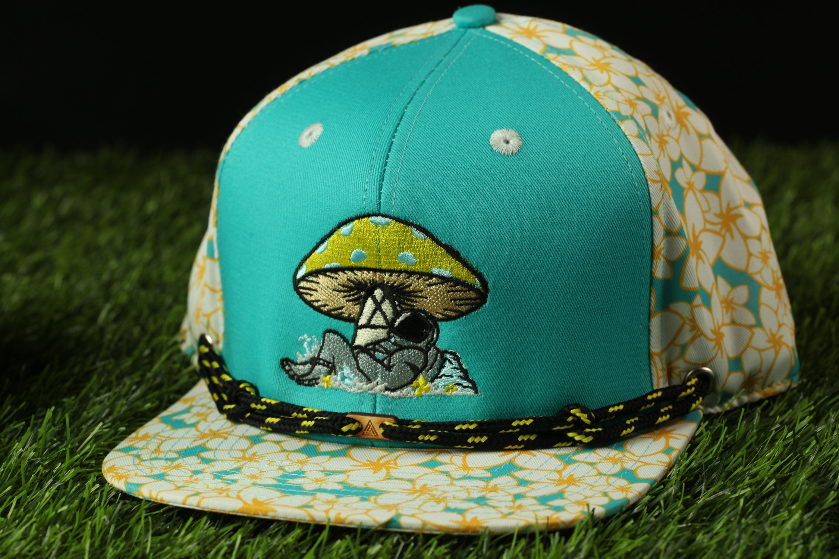 A turquoise baseball cap with a mushroom design and a black and yellow patterned band, placed on a grassy surface.