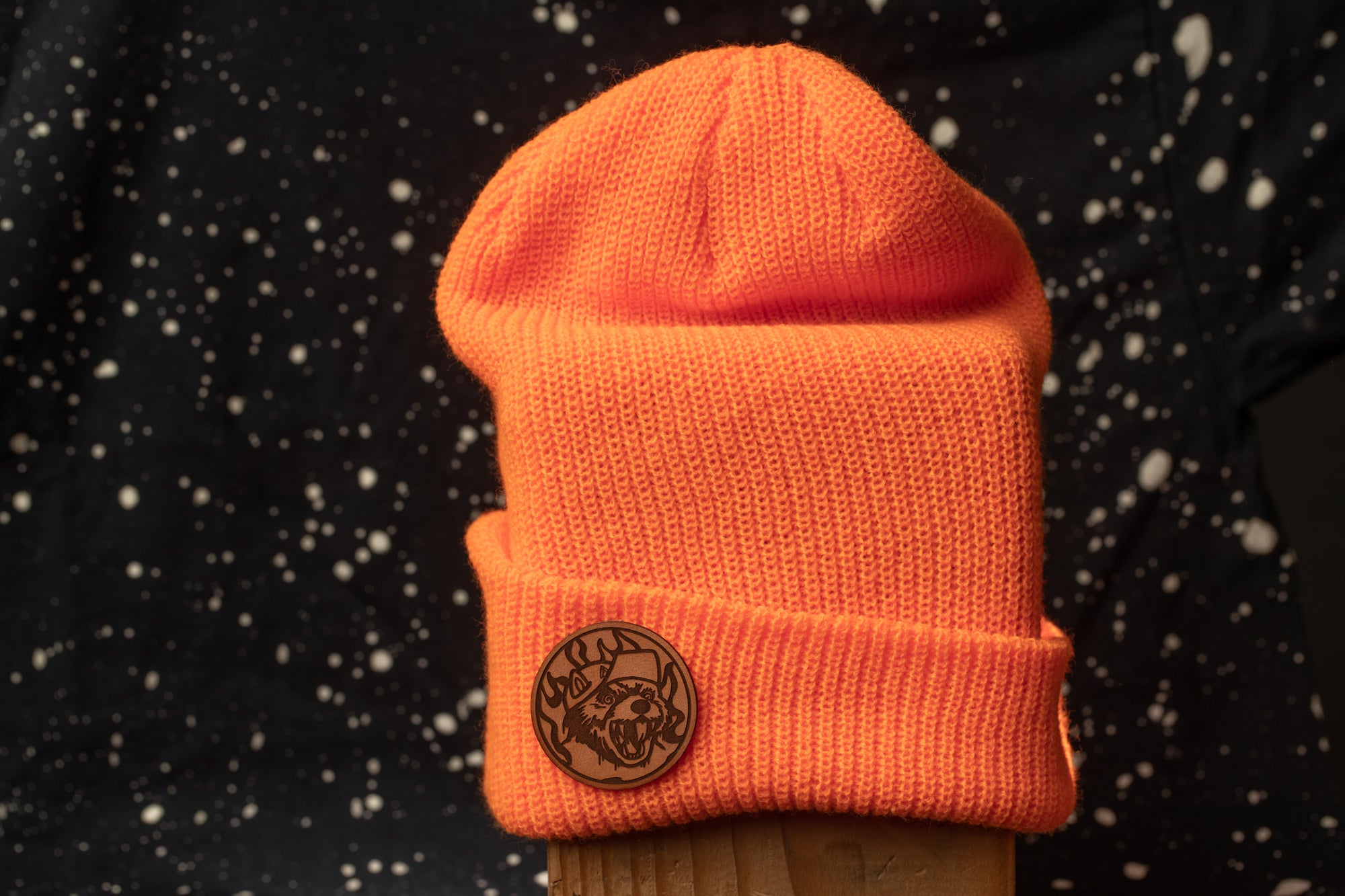 An orange knit beanie with a circular logo on the front, placed against a dark background with white speckles.