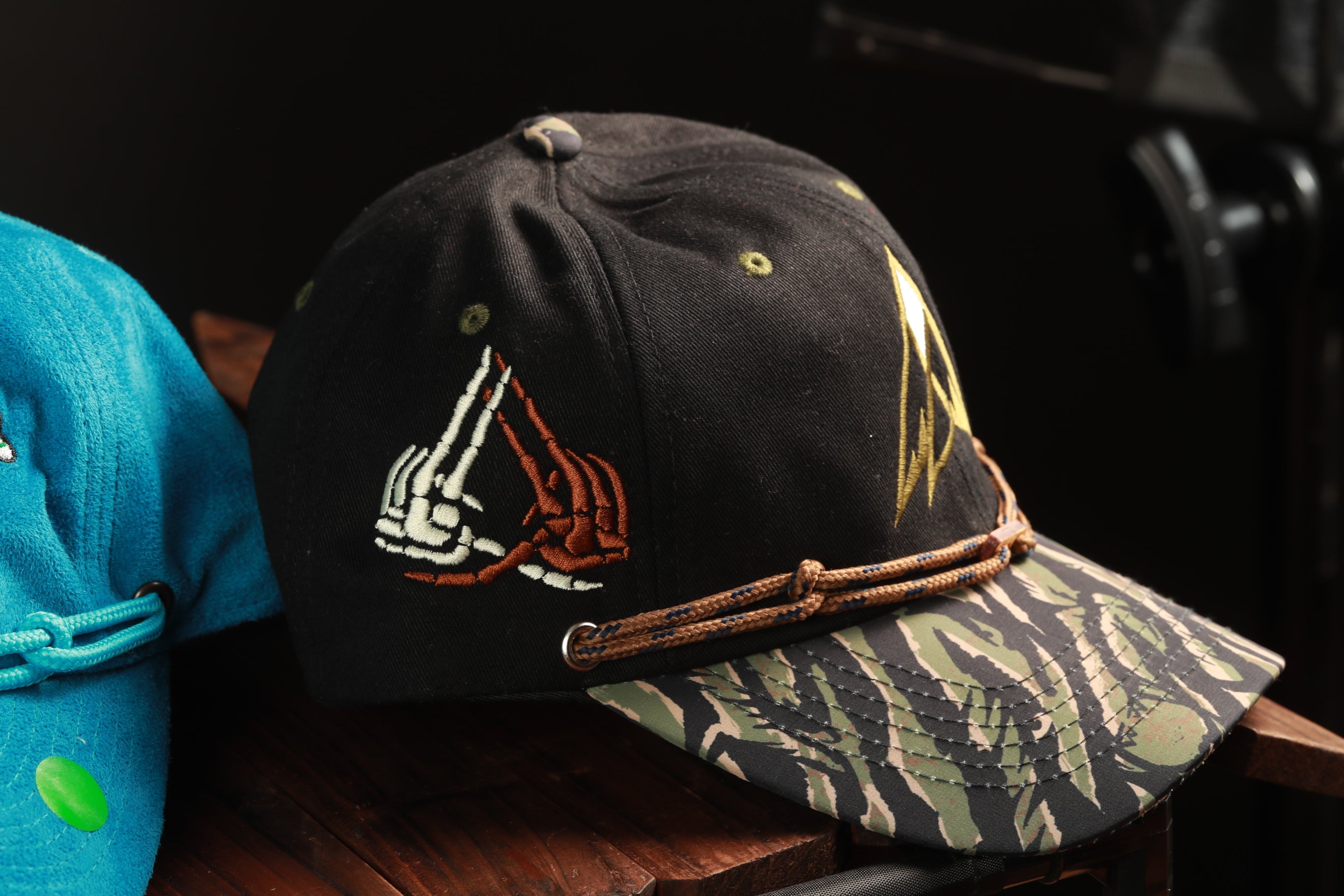 A black baseball cap with a camouflage pattern and a white skull design on the front, sitting on a wooden surface.