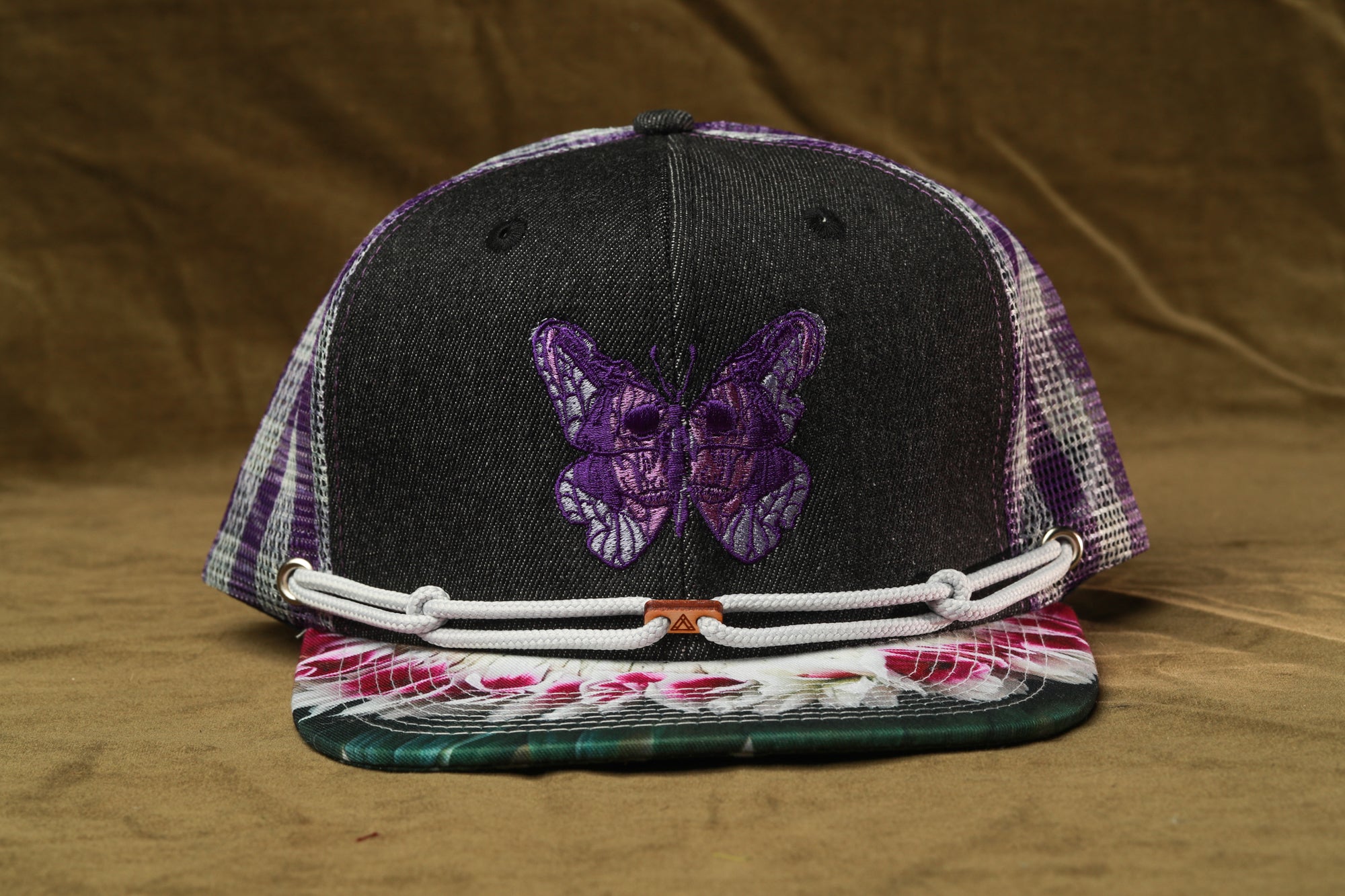 A black baseball cap with a purple butterfly embroidered on it, placed on a brown surface.
