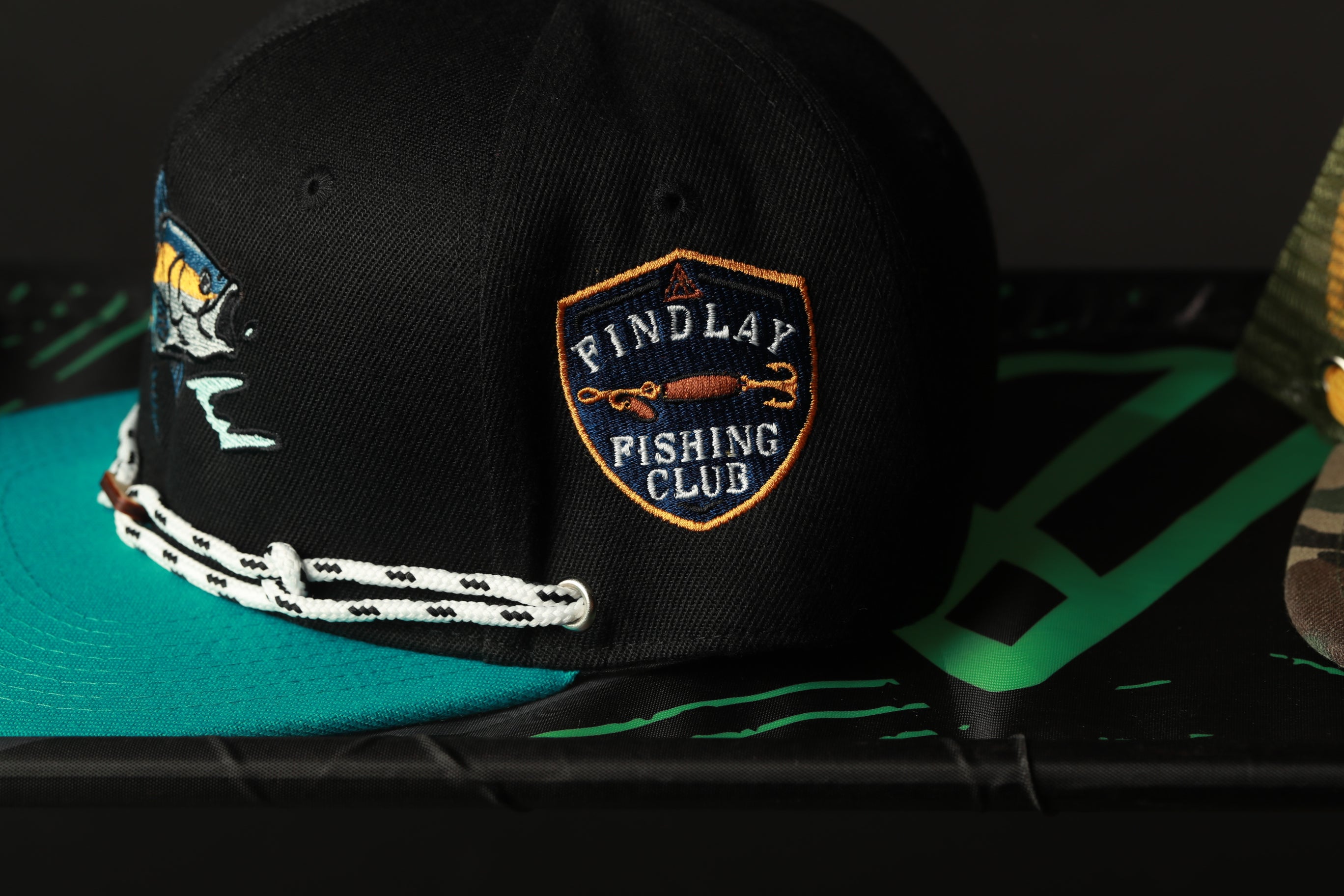 A black baseball cap with a fishing club logo on it, placed on a surface with a teal-colored object.