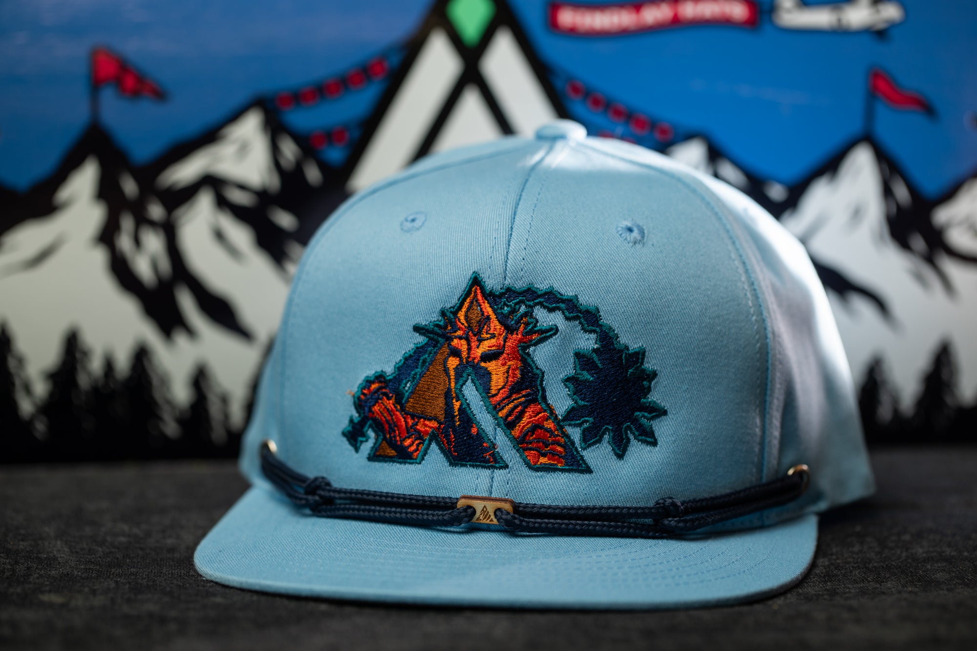 A light blue baseball cap with a colorful, embroidered logo on the front, placed on a surface against a background featuring a mountain landscape with flags.