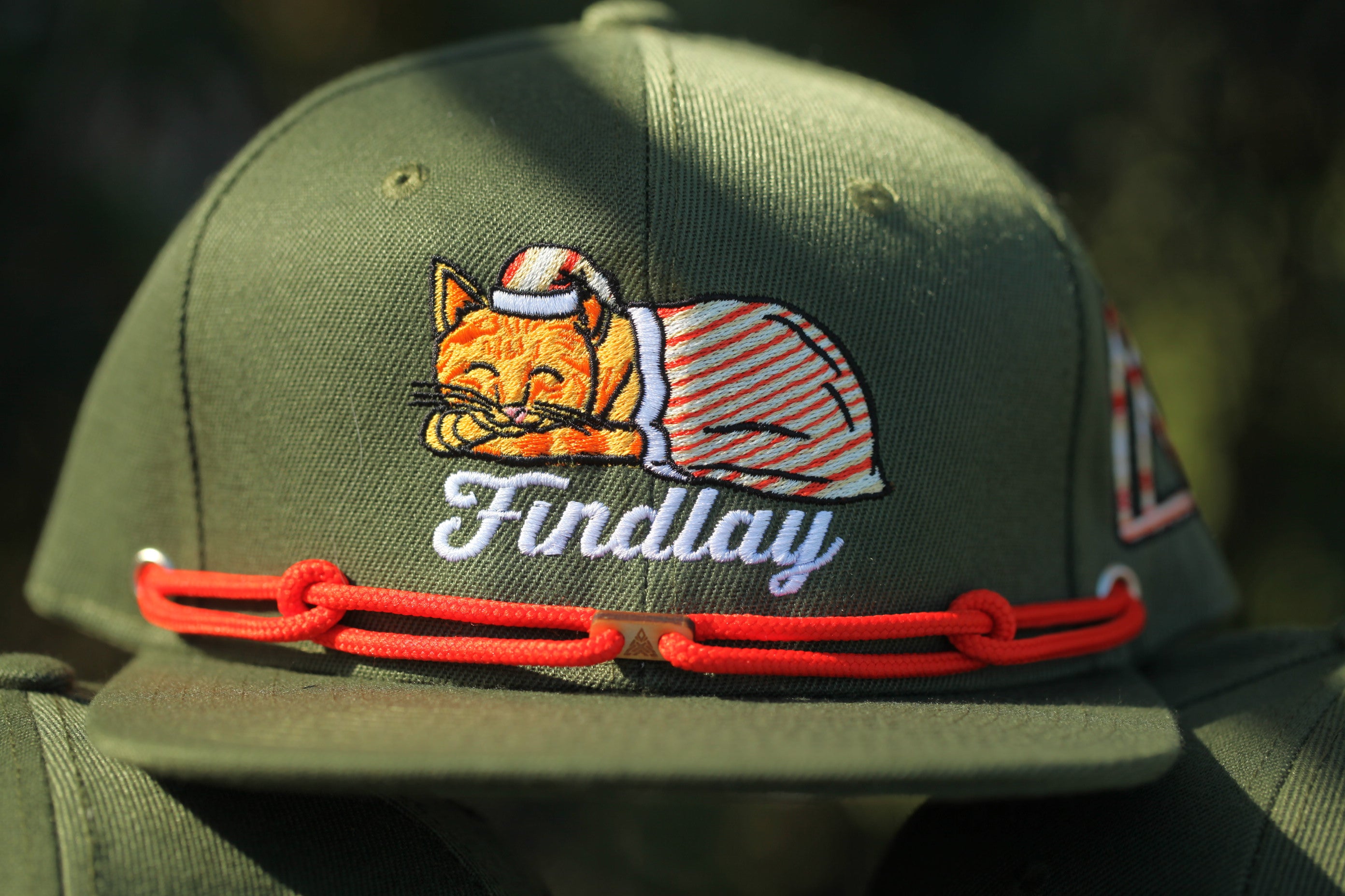 A close-up of a green baseball cap with a cat embroidered on it, wearing a red string.