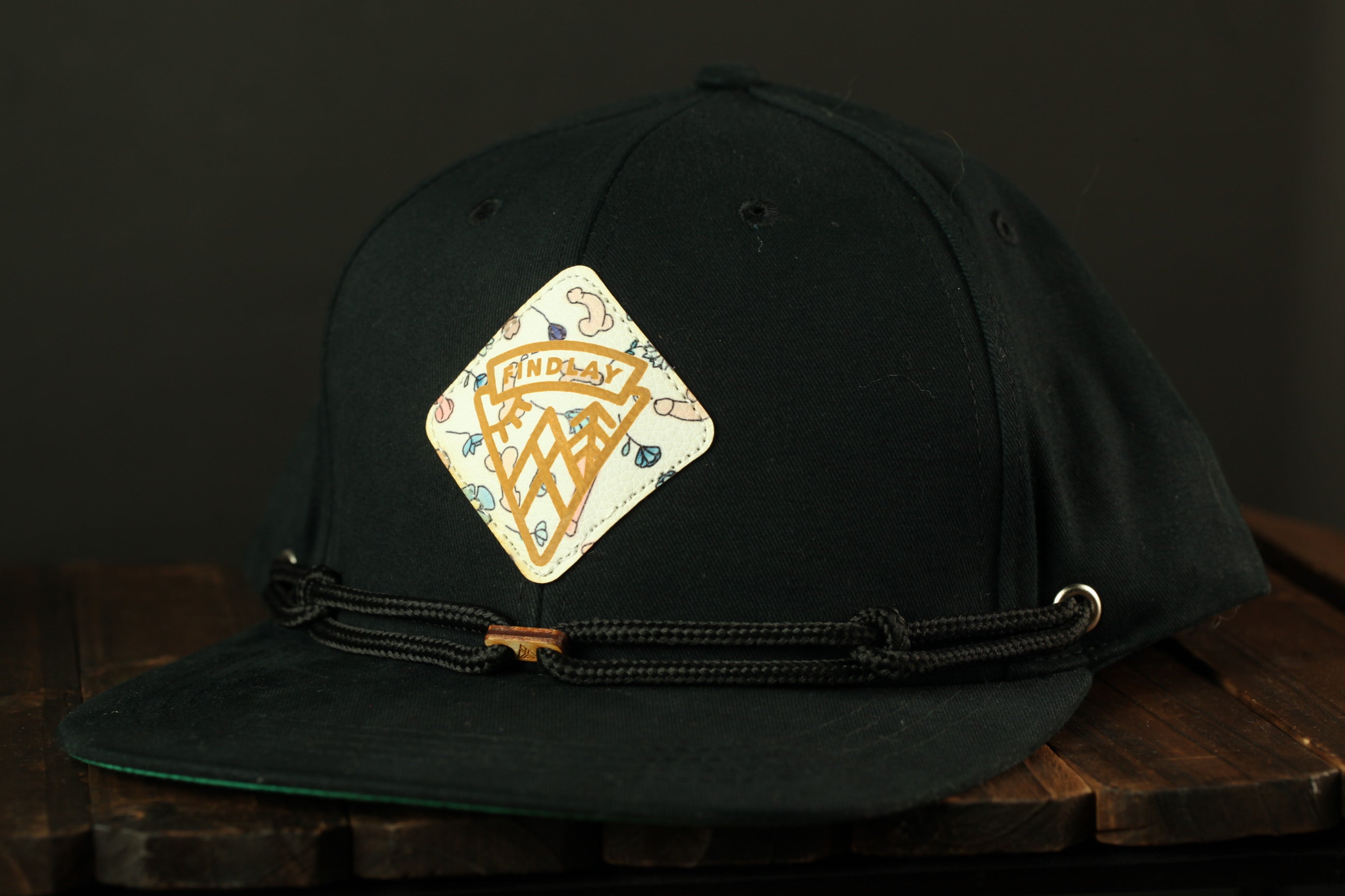 A black baseball cap with a white and gold logo on the front, sitting on a wooden surface.