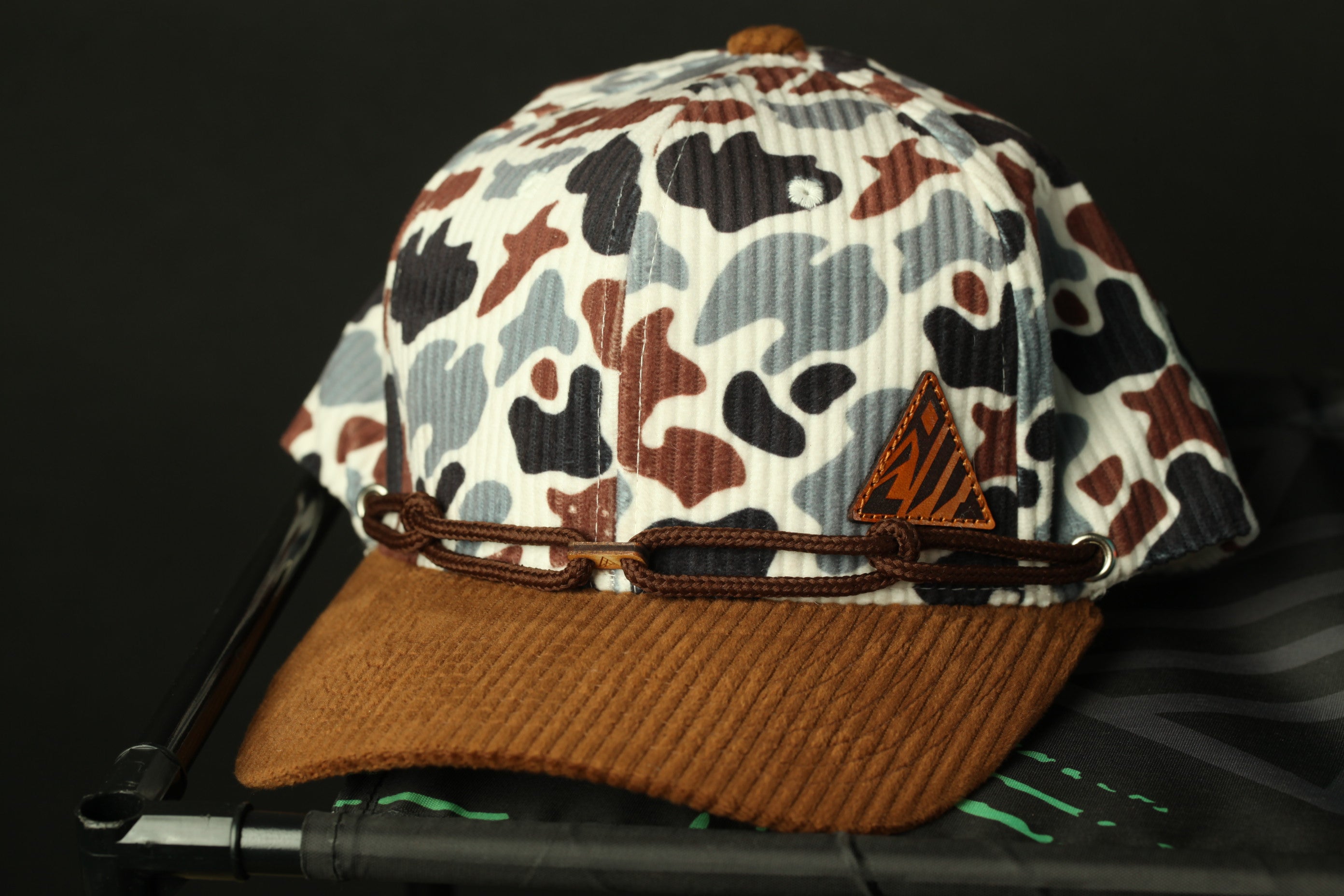 A camouflage-patterned baseball cap with a brown brim and a brown leather band, placed on a black surface.