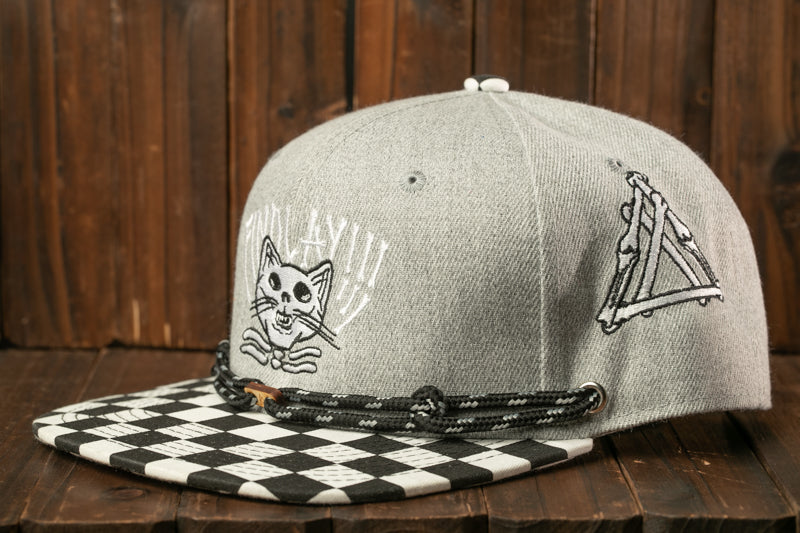 A gray baseball cap with a black and white checkered pattern and a cat design on the front, sitting on a wooden surface.