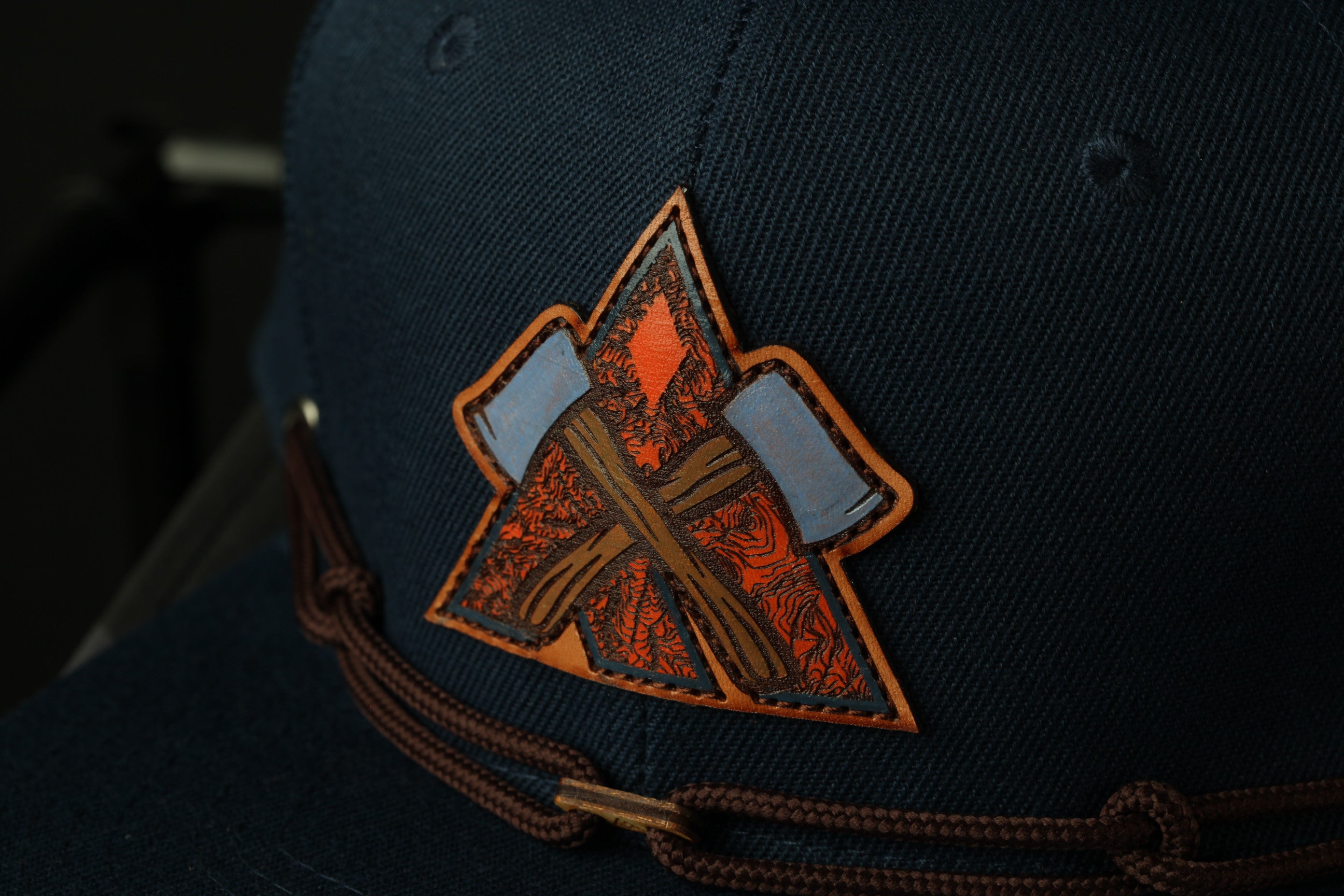 A close-up view of a dark blue baseball cap with a distinctive orange and brown embroidered logo on the front.